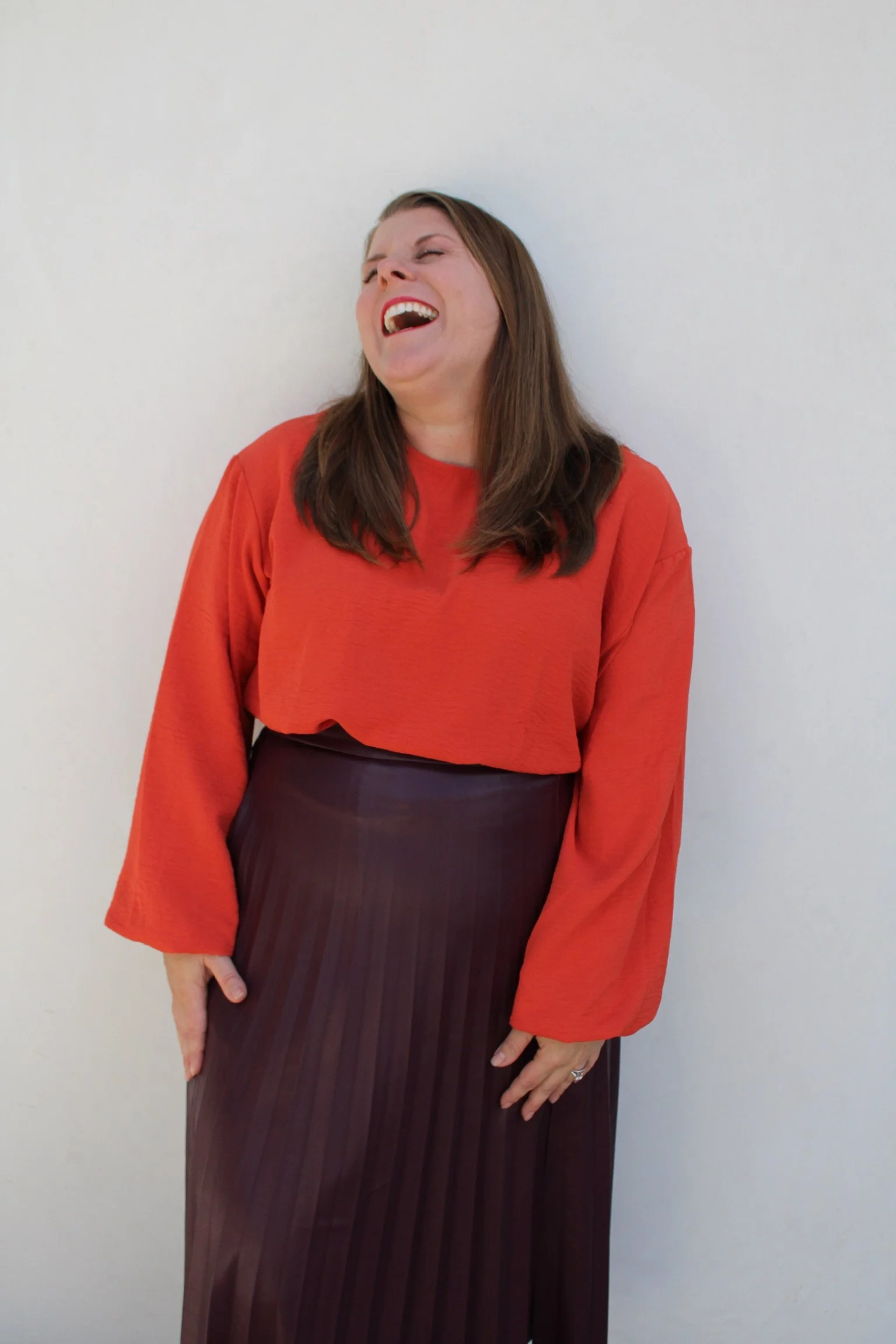 A woman with brown hair laughing, wearing a red long-sleeve top and a dark striped skirt, standing against a plain white wall.