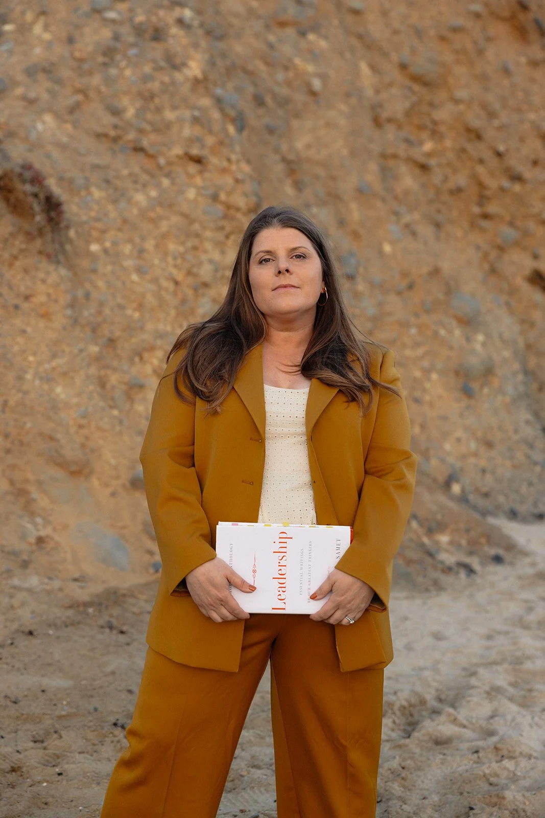 A woman with long brown hair wearing a mustard yellow suit and white shirt, holding a book titled 'Leadership' in front of a dirt and rock background.