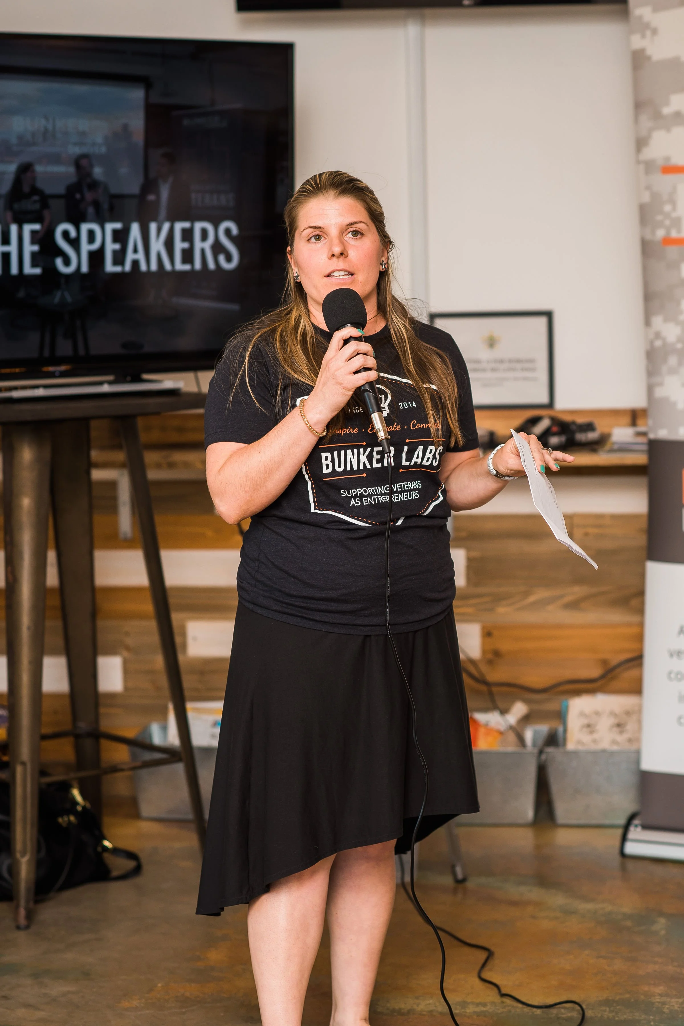 A woman with long hair, wearing a black T-shirt and black skirt, speaking into a microphone in a room with wooden walls and a large screen behind her that displays the words 'THE SPEAKERS'. She is holding a piece of paper in her left hand.