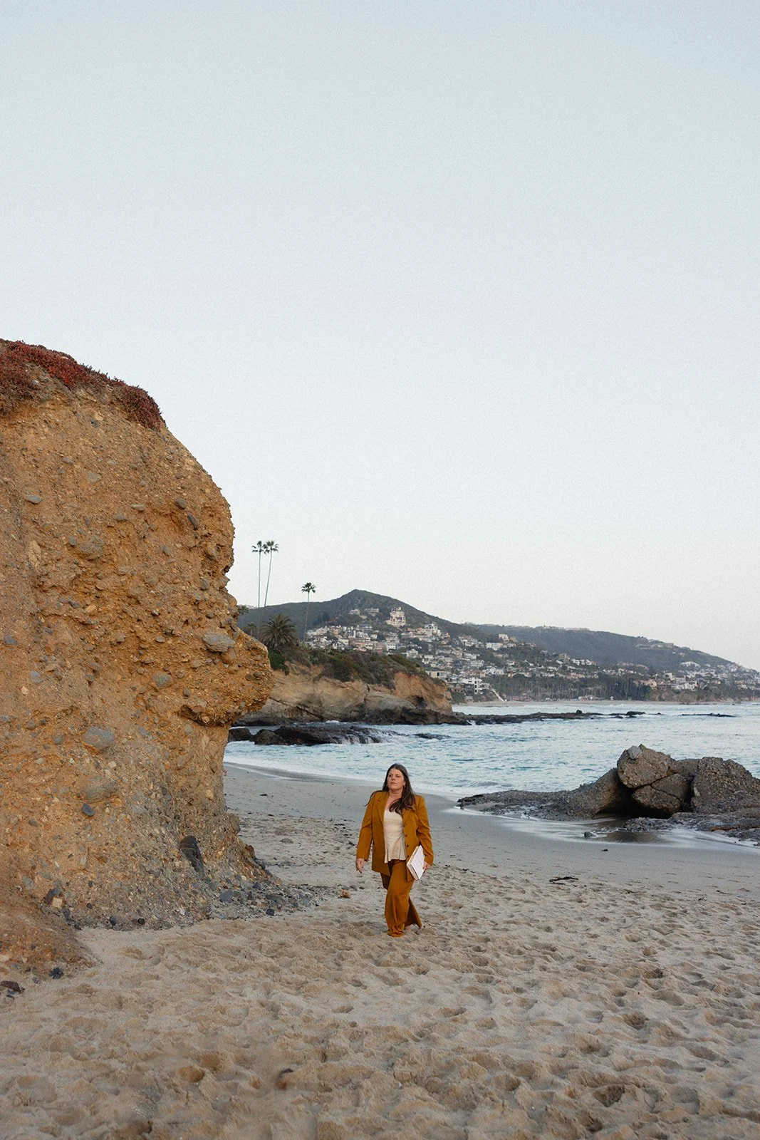 A woman in a mustard-colored suit walking on a sandy beach near a large rocky cliff, with coastal hills and houses in the background.