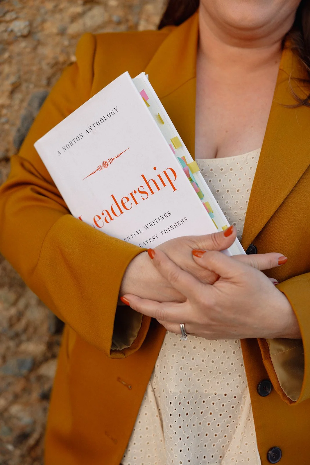 A woman dressed in a mustard blazer is holding a book titled 'Leadership' with colorful dividers and a white cover, standing outdoors near rocks.