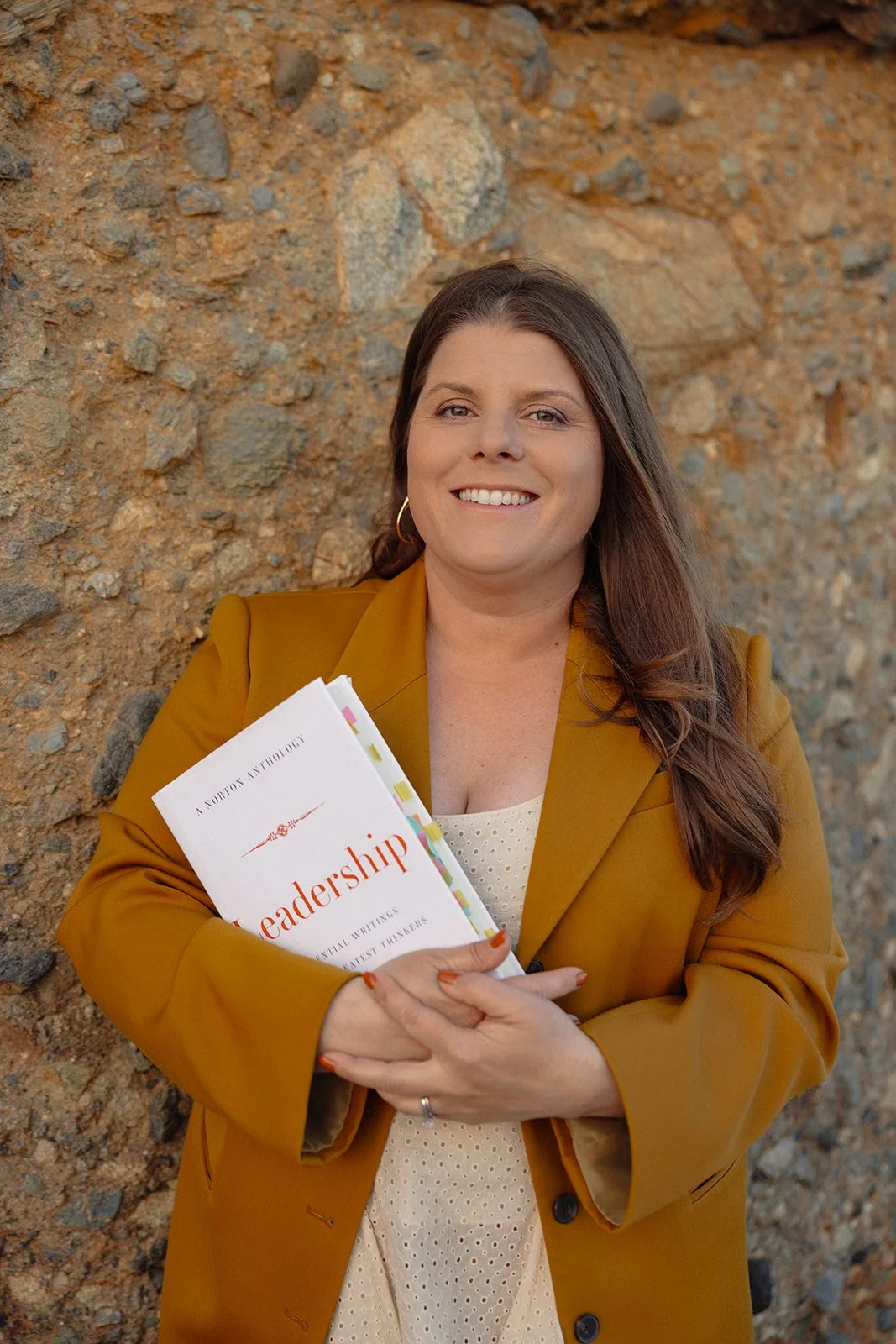 Smiling woman with long brown hair, wearing a mustard blazer and holding a book titled 'Leadership' in front of a rocky background.