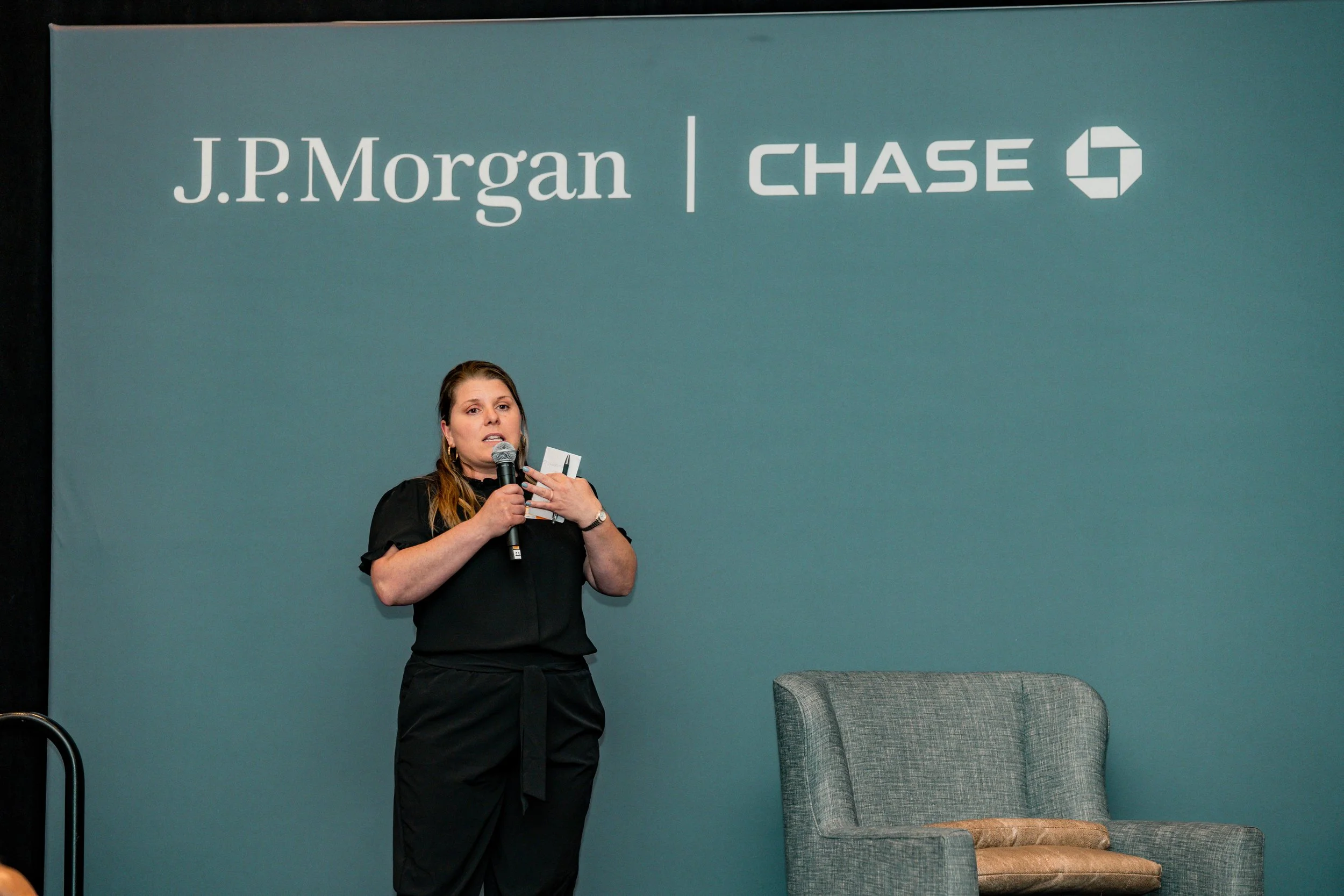 A woman speaking into a microphone during a presentation, standing in front of a blue wall with the logos of J.P. Morgan and Chase.