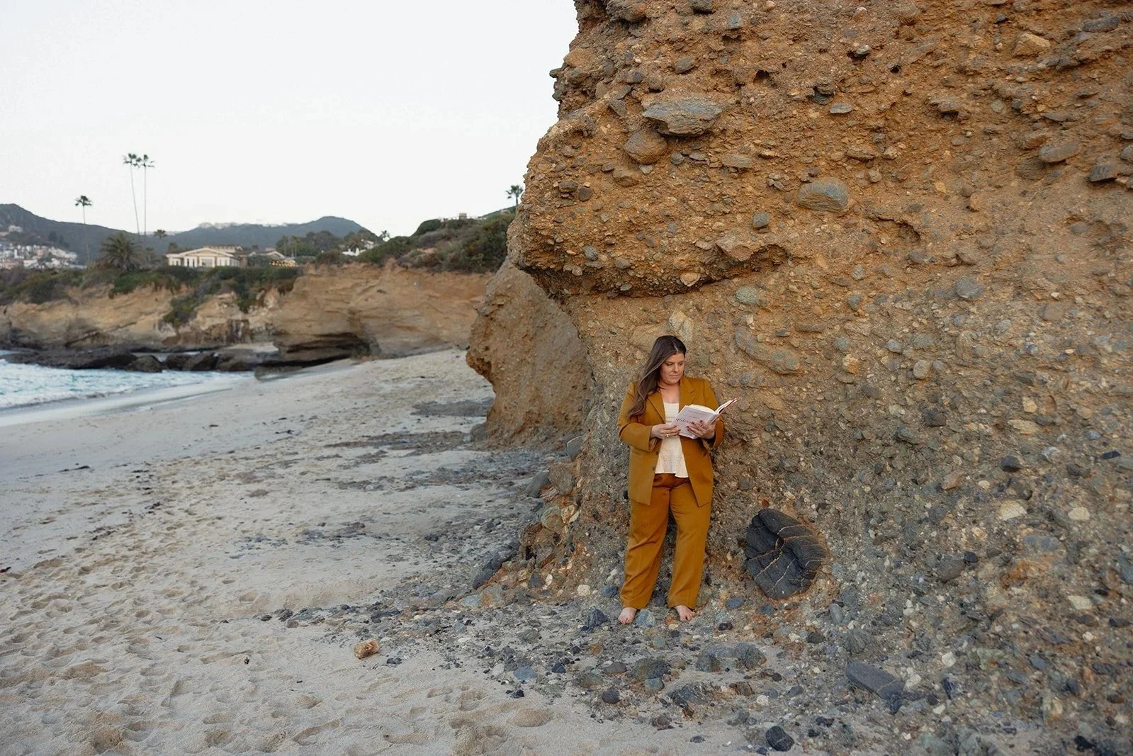 A woman in mustard-colored suit reading a book while standing barefoot against a large rocky cliff on a beach with sandy shore, ocean, and distant cliffs and houses.