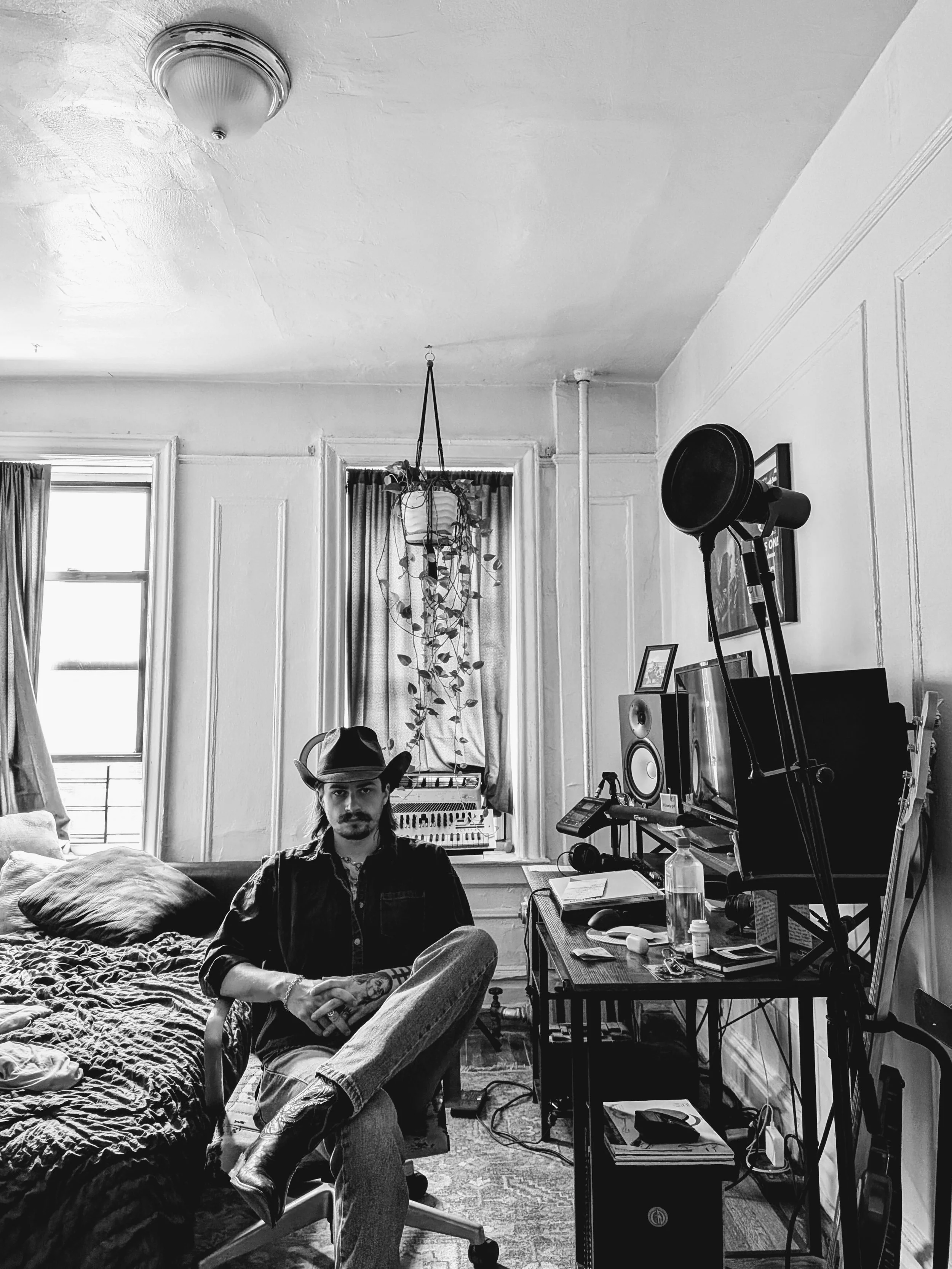 Black and white photo of a man with long hair, a beard, wearing a cowboy hat, sitting on a chair in a room filled with music equipment, including a computer, speakers, and musical instruments. The room has large windows with curtains, and a plant hanging from the ceiling.