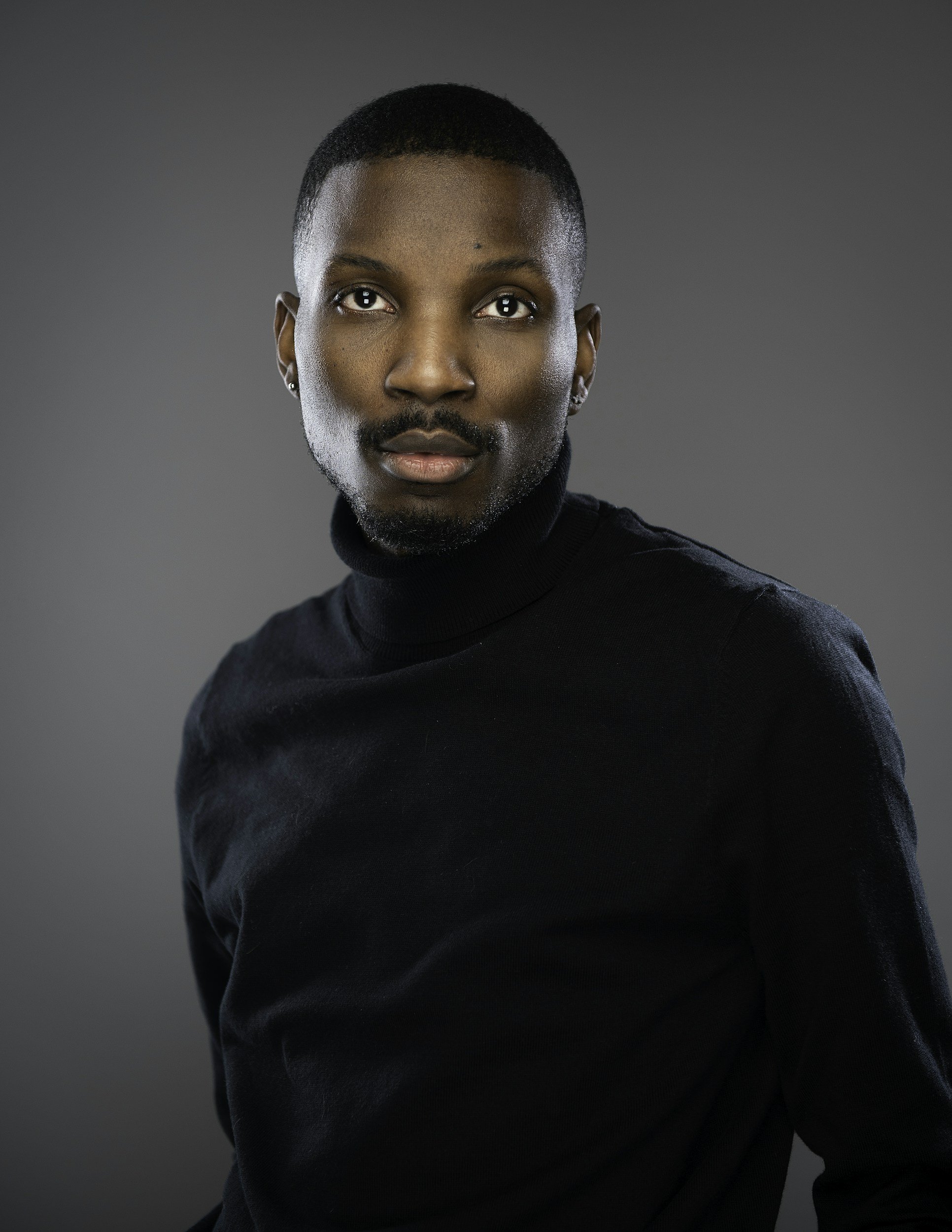 A portrait of an Black woman with short hair, wearing a black turtleneck sweater, against a gray background.