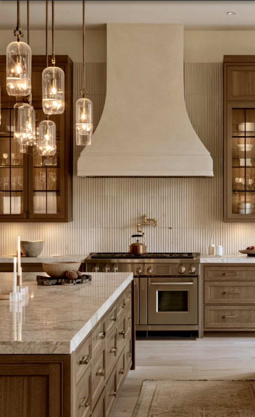 Warm transitional kitchen designed by Indigo Day Designs, featuring a fluted backsplash, glass-front cabinetry, statement lighting, and a sculptural hood.
