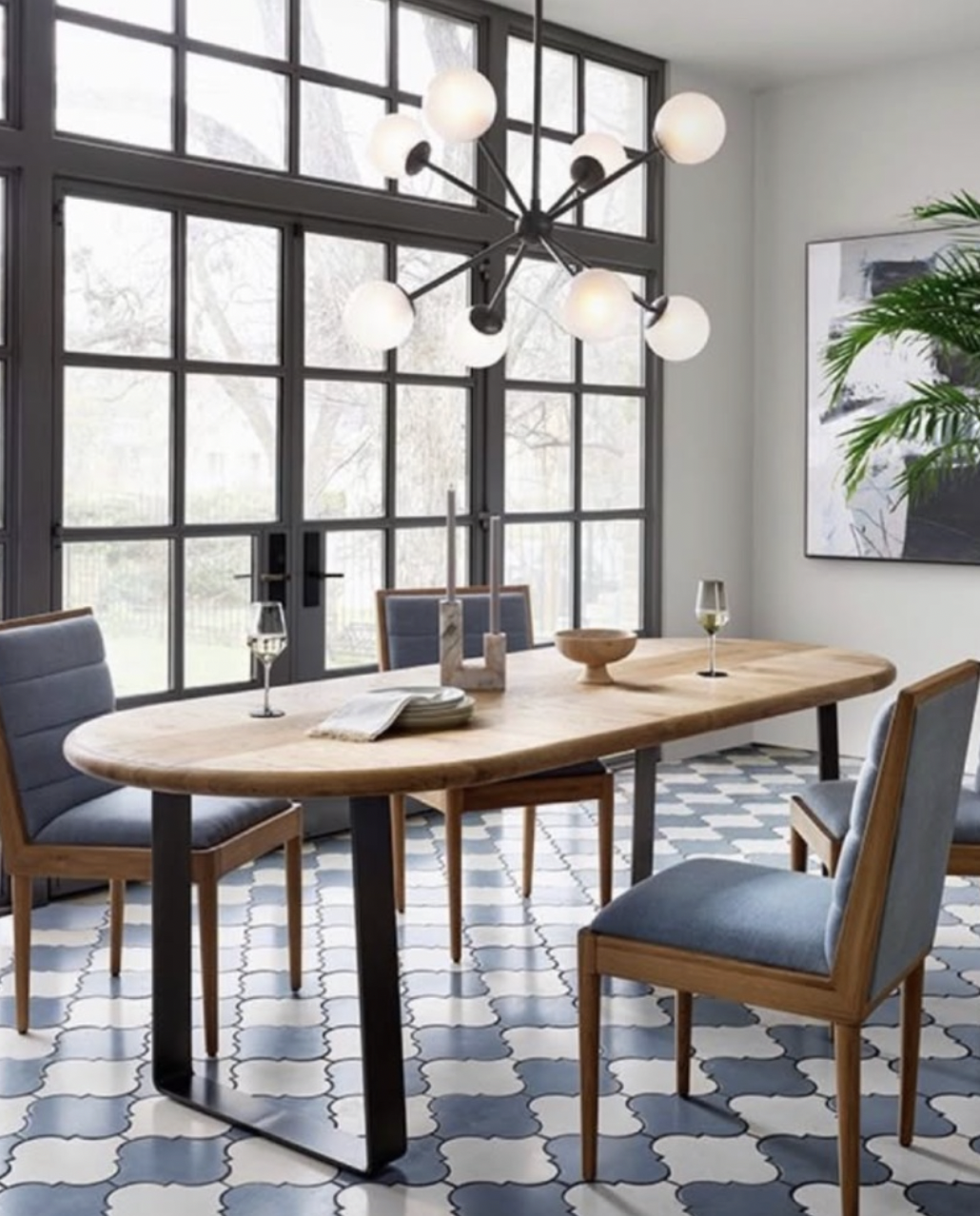 A modern dining room with a wooden table, four cushioned chairs, large windows, a black and white geometric patterned tile floor, a contemporary chandelier, candles, books, and wine glasses on the table, and a large plant and artwork on the wall.