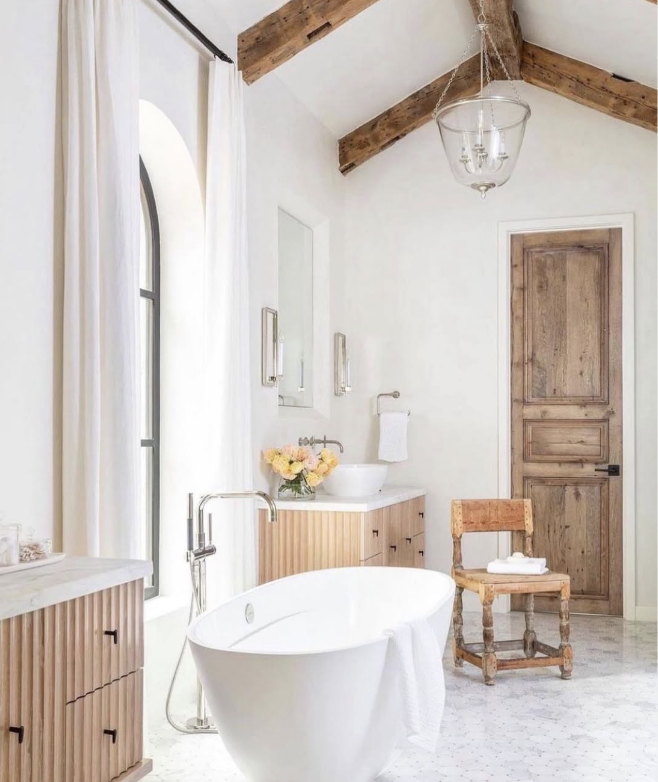 Bathroom with modern freestanding bathtub, wooden cabinets, a small sink, a vase with flowers, a wooden chair, a door, and a window with white curtains, wooden beams overhead, and a hanging light fixture.