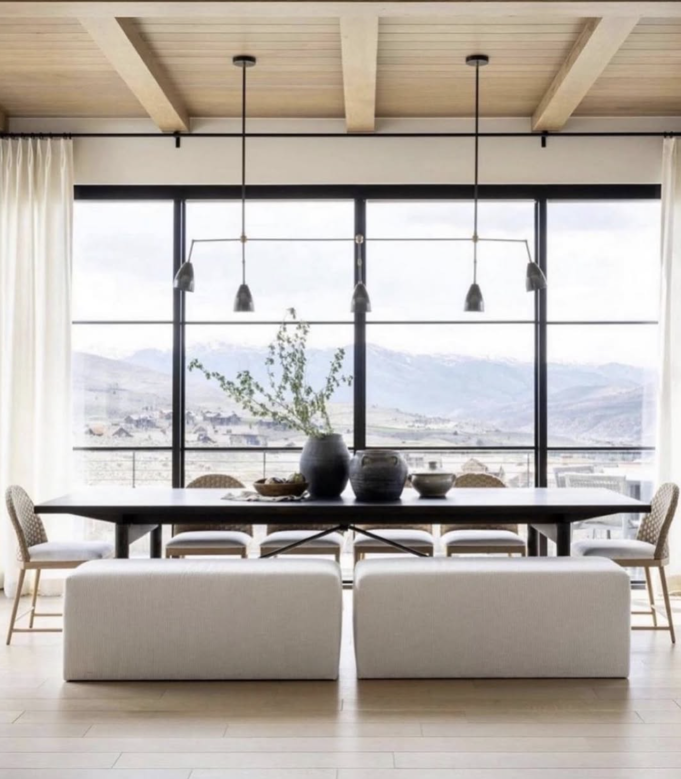 Modern dining room with large floor-to-ceiling windows, white curtains, wooden ceiling beams, and a mountain view, featuring a black dining table with decorative vases and chairs with white cushions.