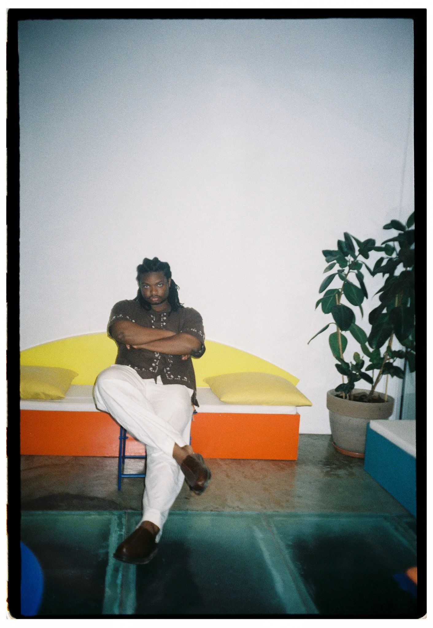 A man with dreadlocks wearing a brown shirt and white pants sitting on a colorful bench with yellow pillows, crossing his arms and leg, in a room with a blank wall, a potted plant, and colorful furniture.
