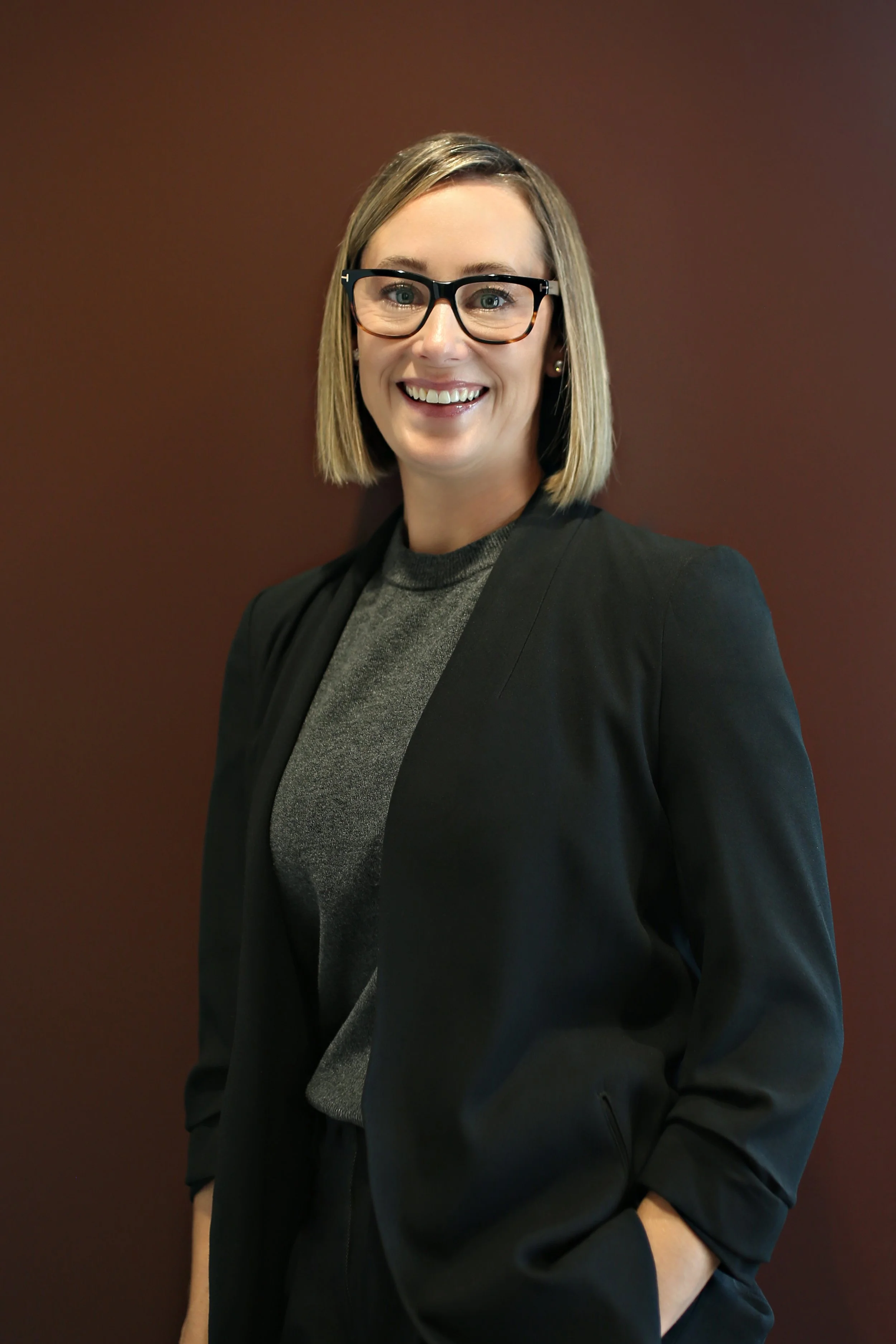 A woman with blonde hair, glasses, and earrings, wearing a grey top and black blazer, smiling in front of a plain dark brown background.