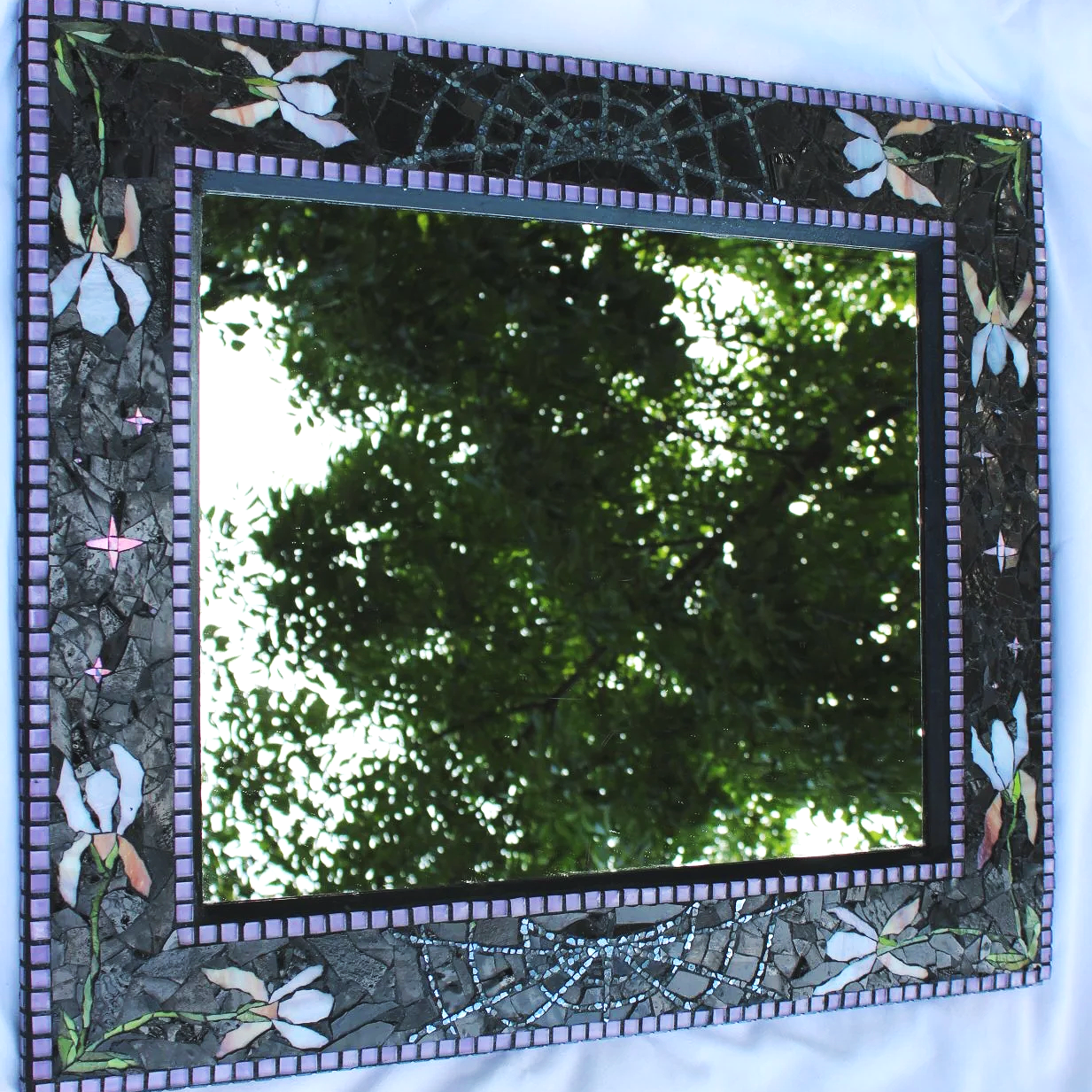 A rectangular mosaic mirror with a decorative border featuring floral and spider web patterns, reflecting green leafy trees and sunlight.