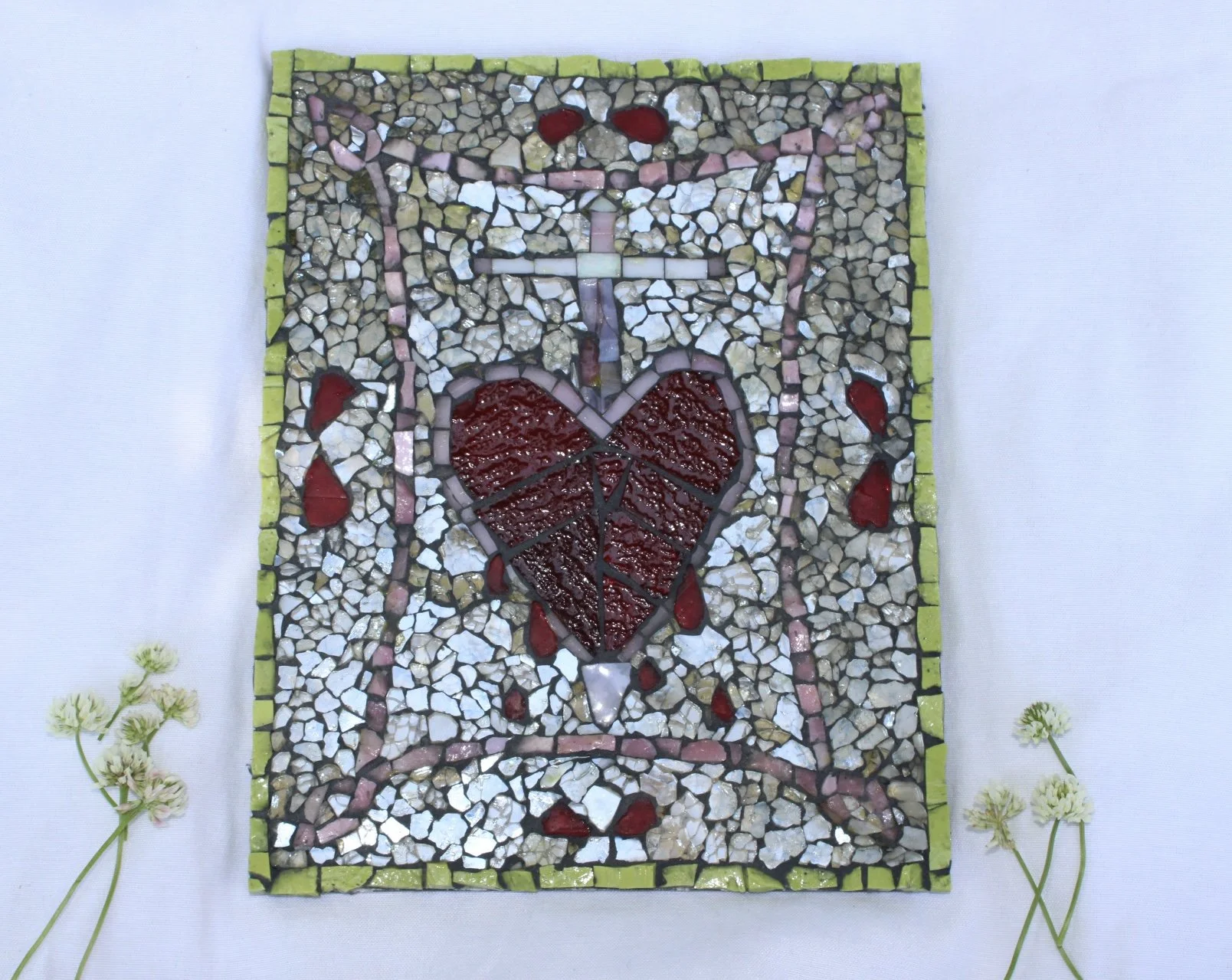 Mosaic art piece depicting a broken heart with a sword through it, dripping blood, framed by small green tiles, with a white glass mosaic background.