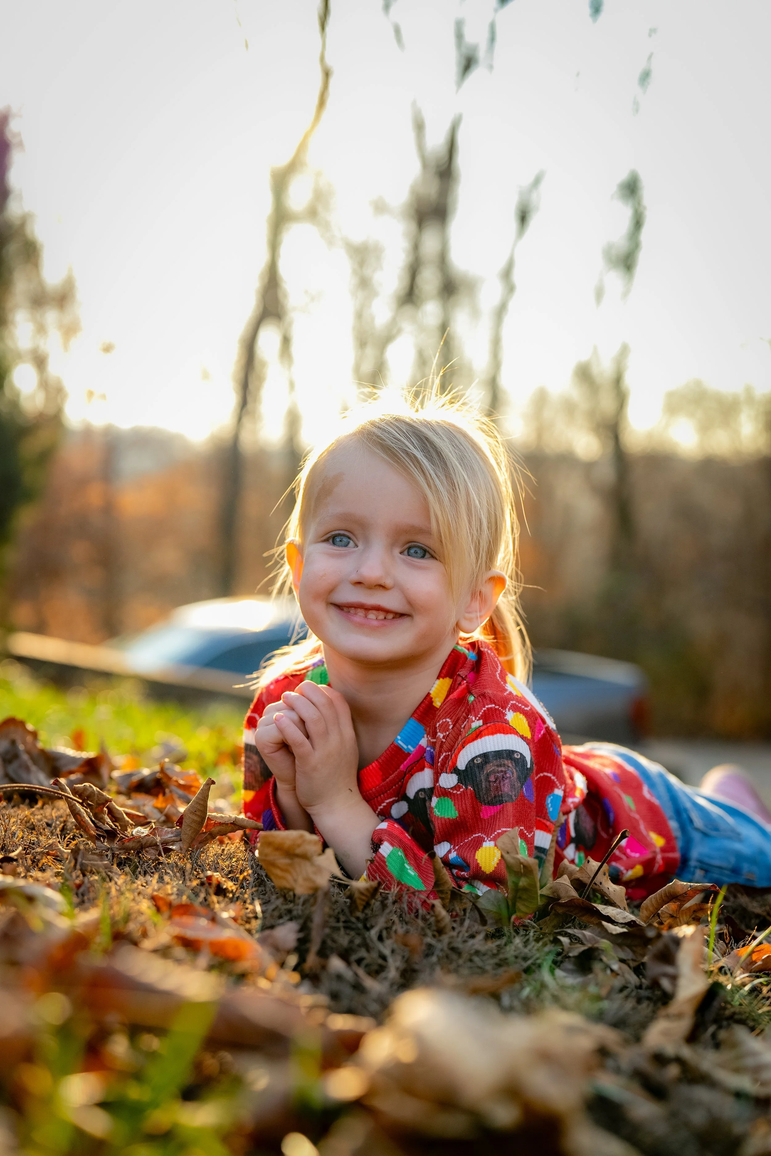 Young girl with blonde hair and blue eyes lying on her stomach outdoors during autumn, smiling, surrounded by fallen leaves with trees and sunshine in the background.