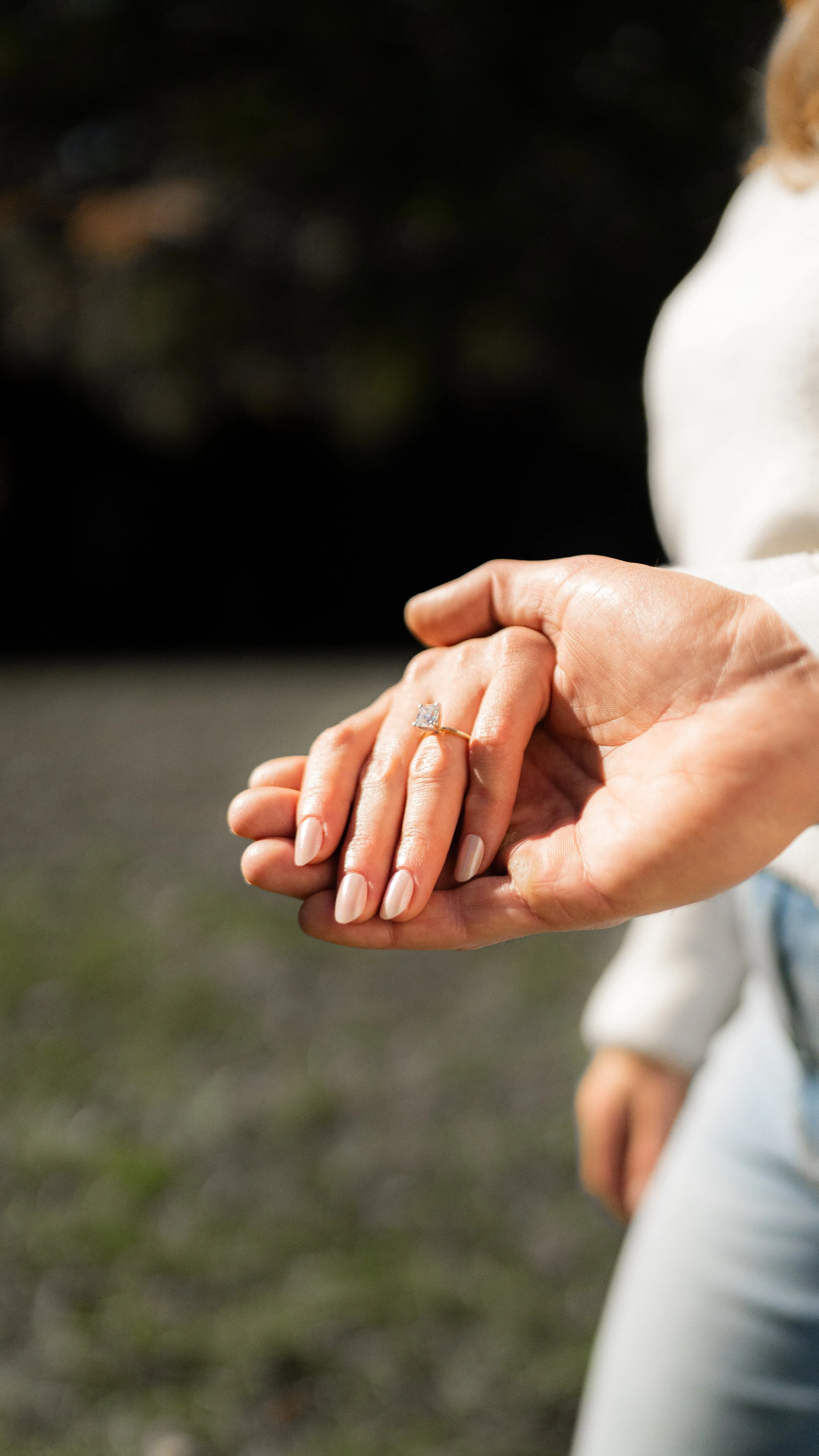 Close-up of a woman's hand with white nail polish, wearing a diamond engagement ring, being held by a man's hand, outdoors with a blurred natural background.