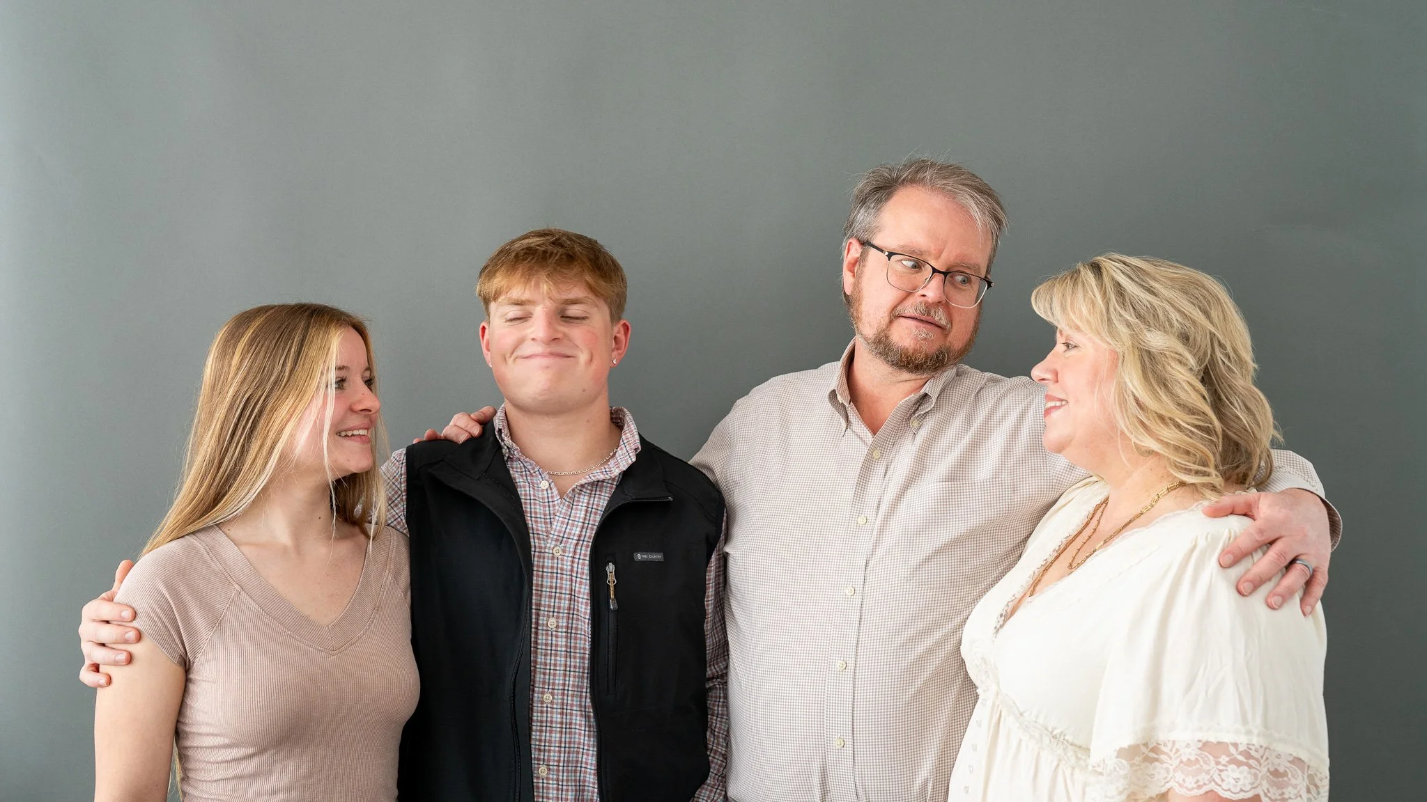 Family picture of five people standing close together against a plain grey background, smiling and looking at each other.