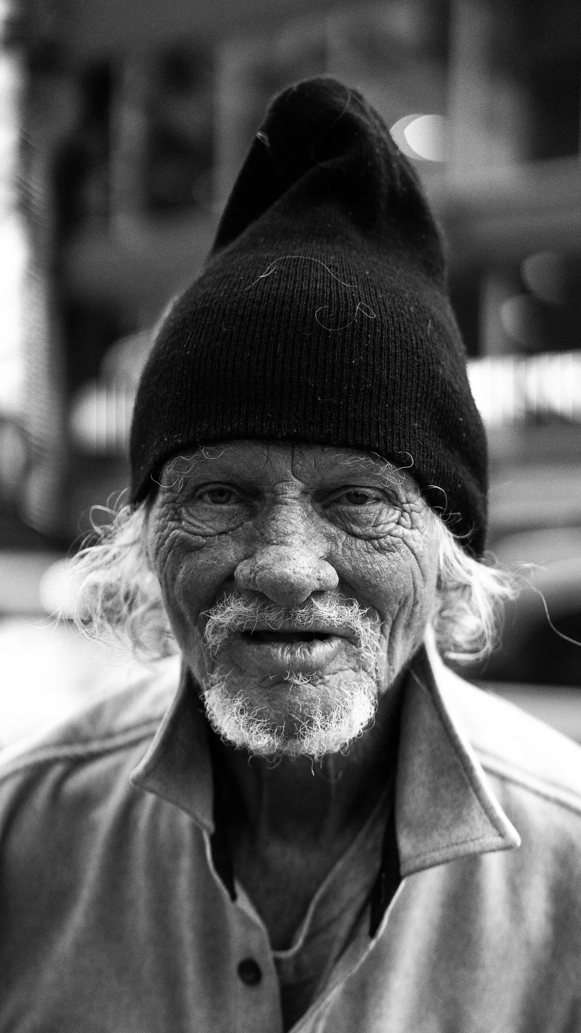A black and white portrait of an elderly man with a furrowed face, white hair, and a beard, wearing a knit cap and a collared jacket outdoors.