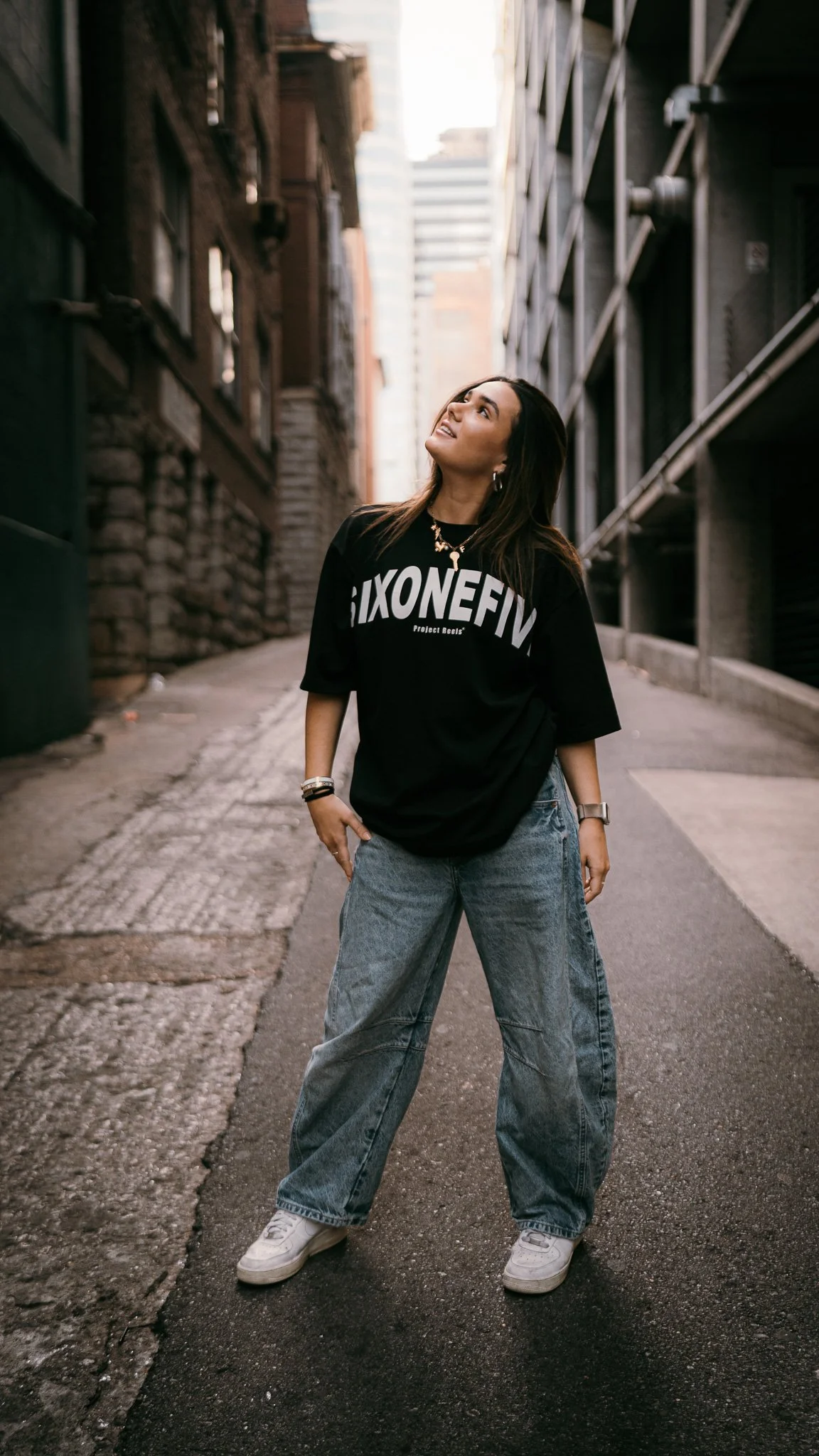 A young woman standing in an alleyway between tall buildings, wearing a black oversized T-shirt with text, loose jeans, and white sneakers, looking upward with a smile.