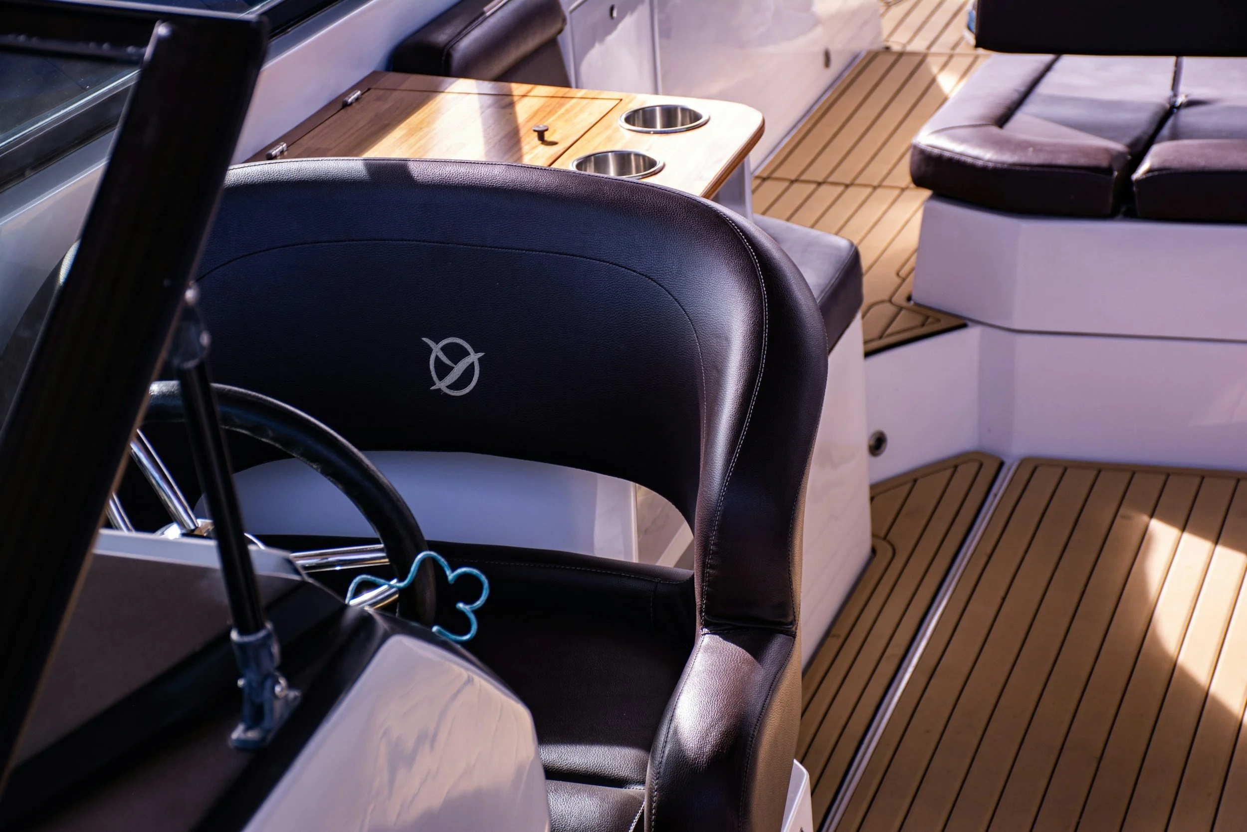 Close-up of boat interior with black leather seats, a wooden table with two cup holders, and a cushioned bench seat with beige cushions on a wooden deck.