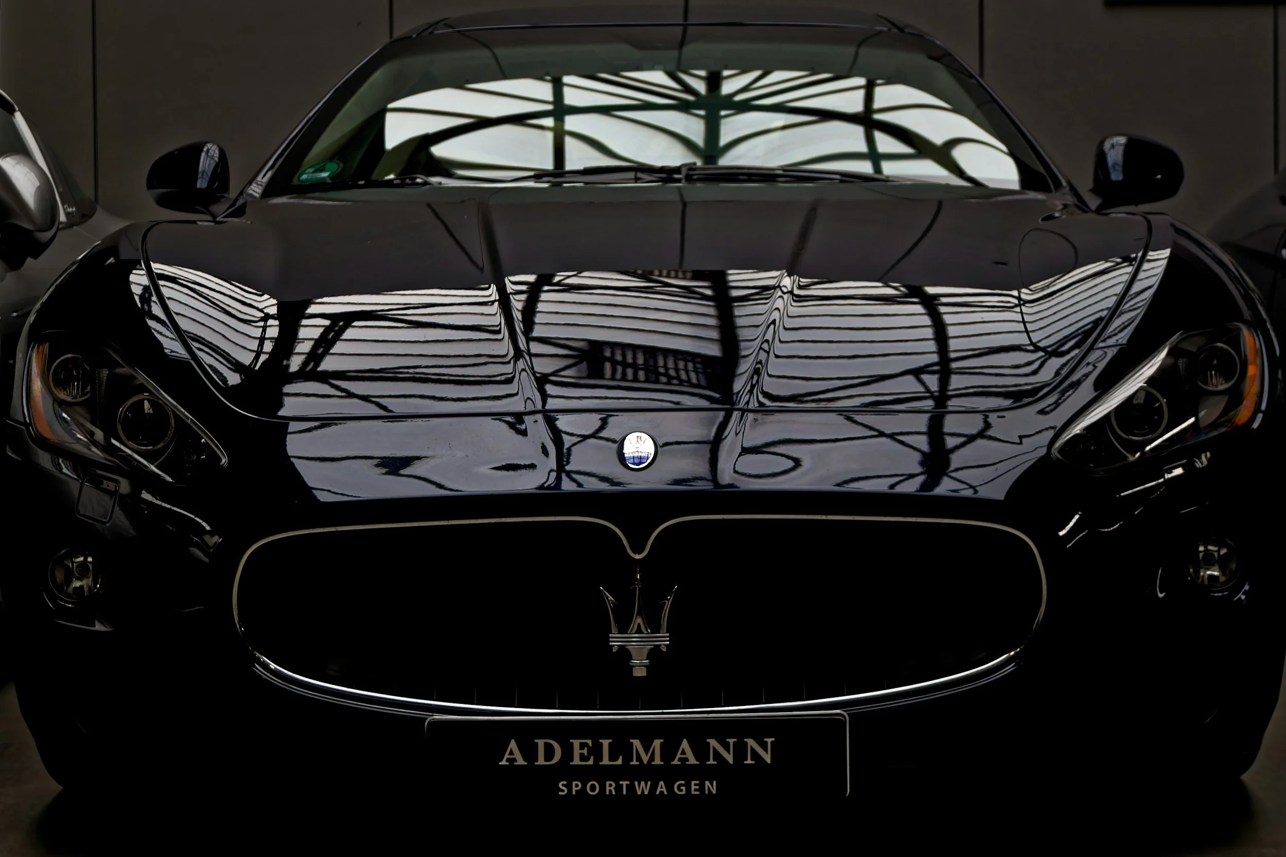 A sleek black sports car with reflective surface, showcasing intricate reflections on its hood, positioned indoors with minimal surrounding objects and a license plate reading 'ADELANN'. - Concours Grade