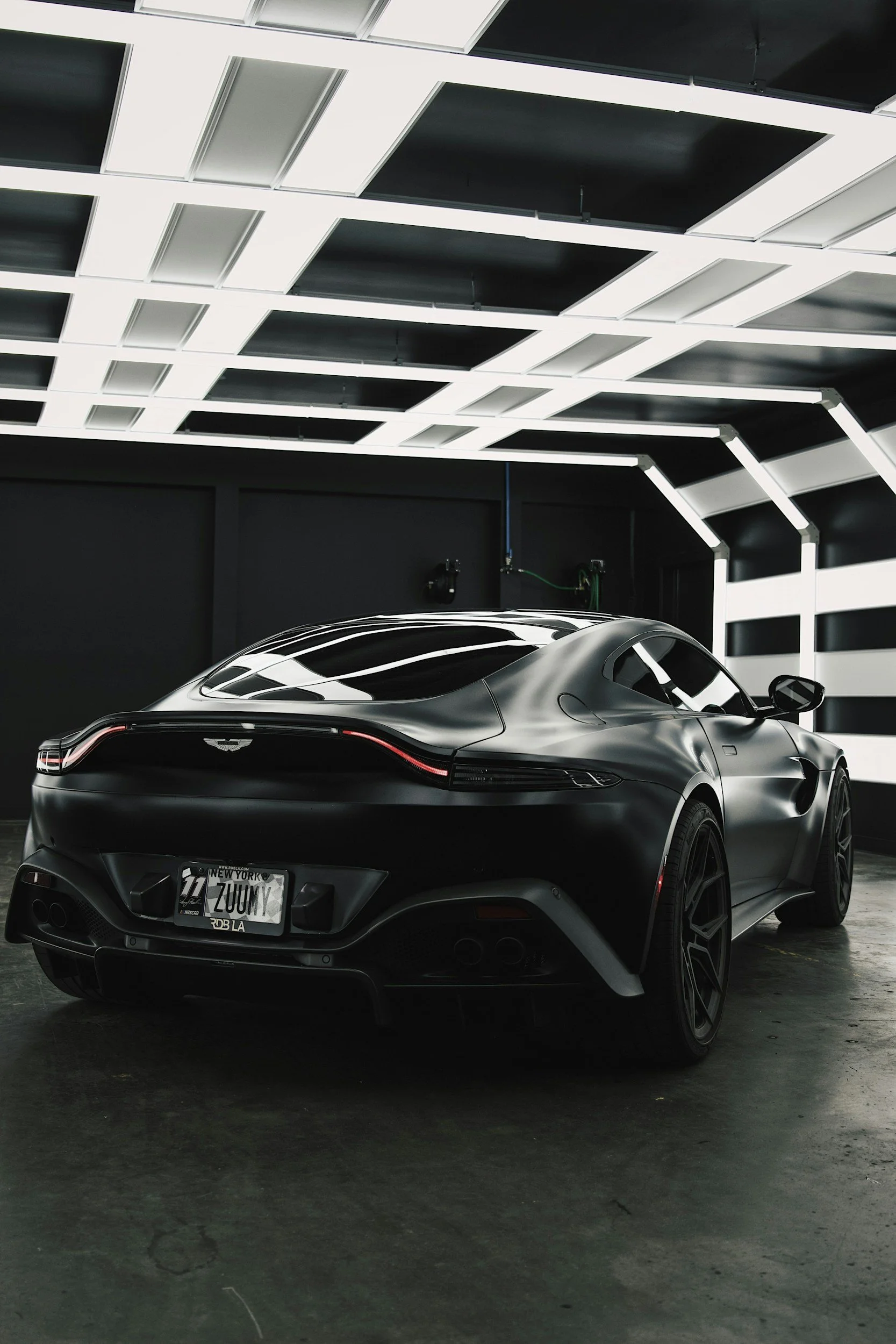 A sleek, black sports car parked indoors under a modern, illuminated ceiling with geometric patterns. The car has a distinctive, futuristic design with a low profile and tinted windows.