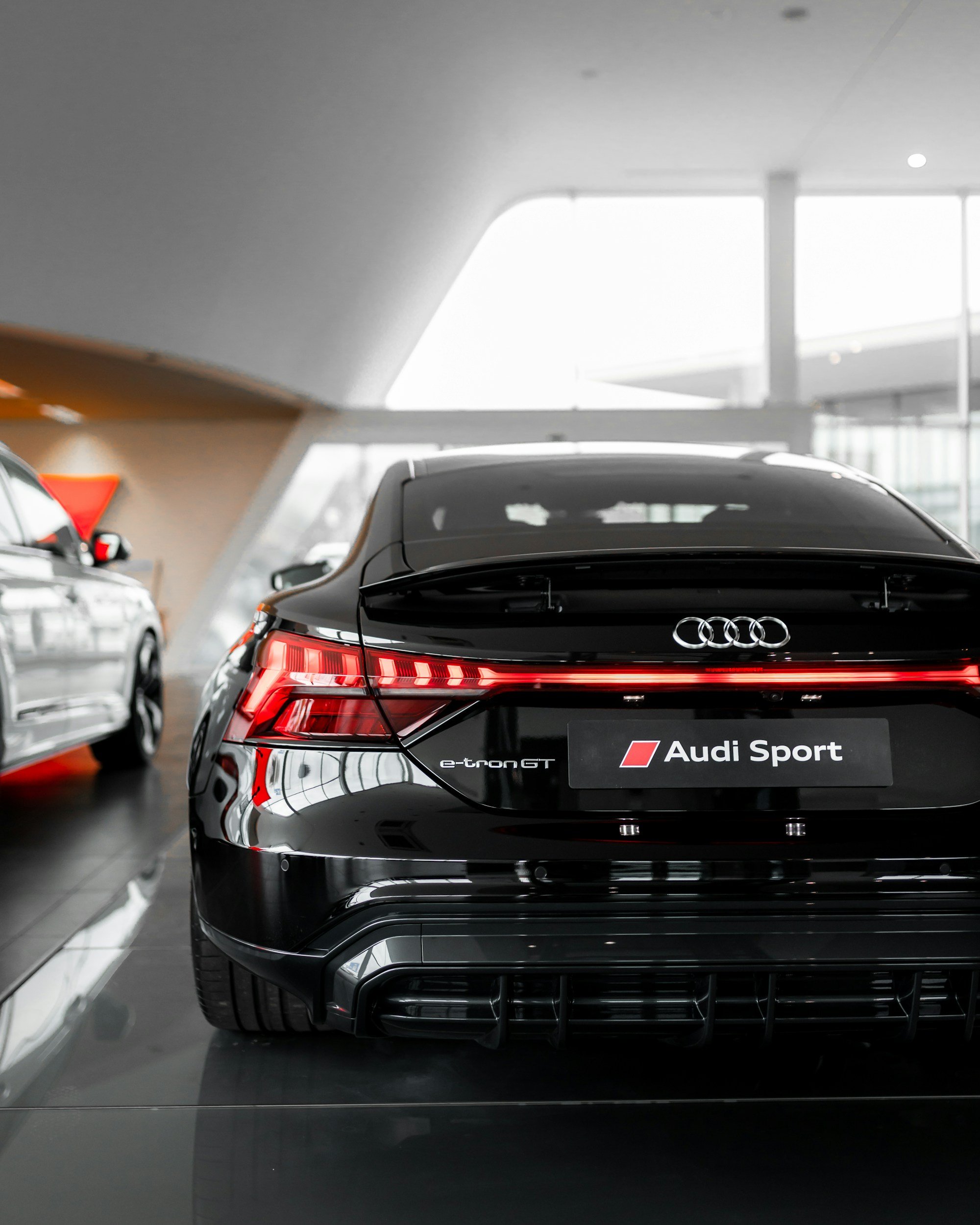 Rear view of a black Audi Sport e-tron GT car in a showroom, with other cars visible on the sides, and a modern building interior in the background.