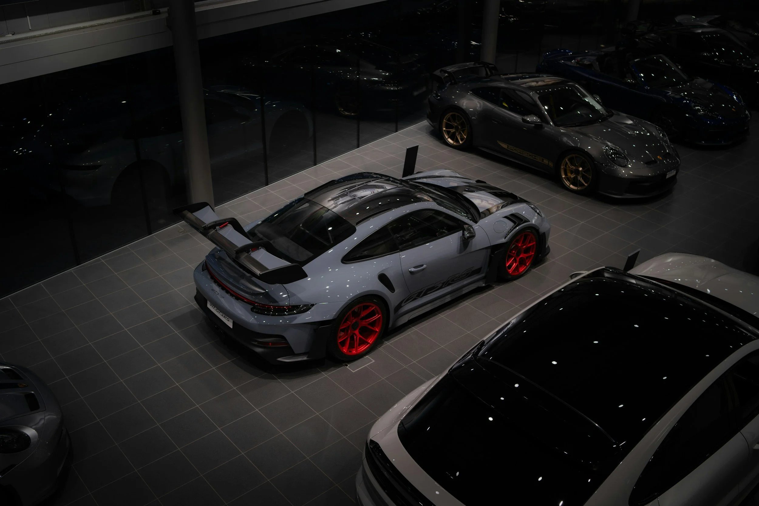 A showroom with luxury sports cars, including a silver Porsche with red wheels, displayed on a tiled floor against a glass wall reflecting other vehicles.