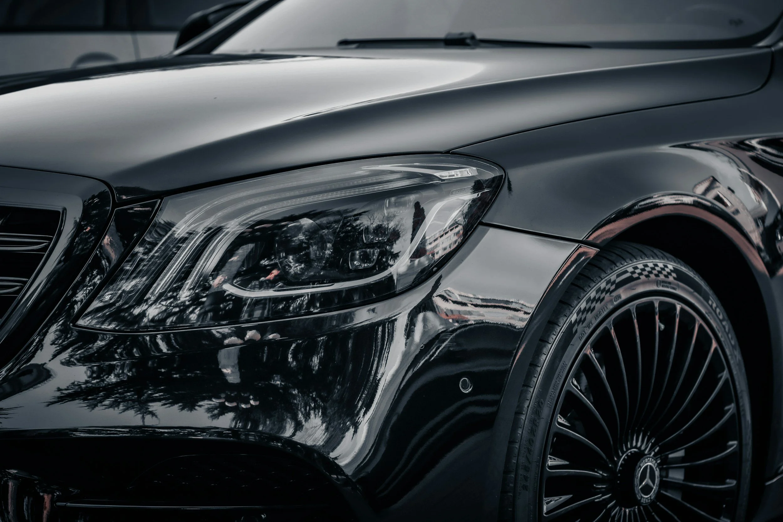 Close-up of a shiny black luxury car, focusing on the front left headlight, fender, and wheel with Mercedes-Benz logo, reflecting nearby trees and cloudy sky.