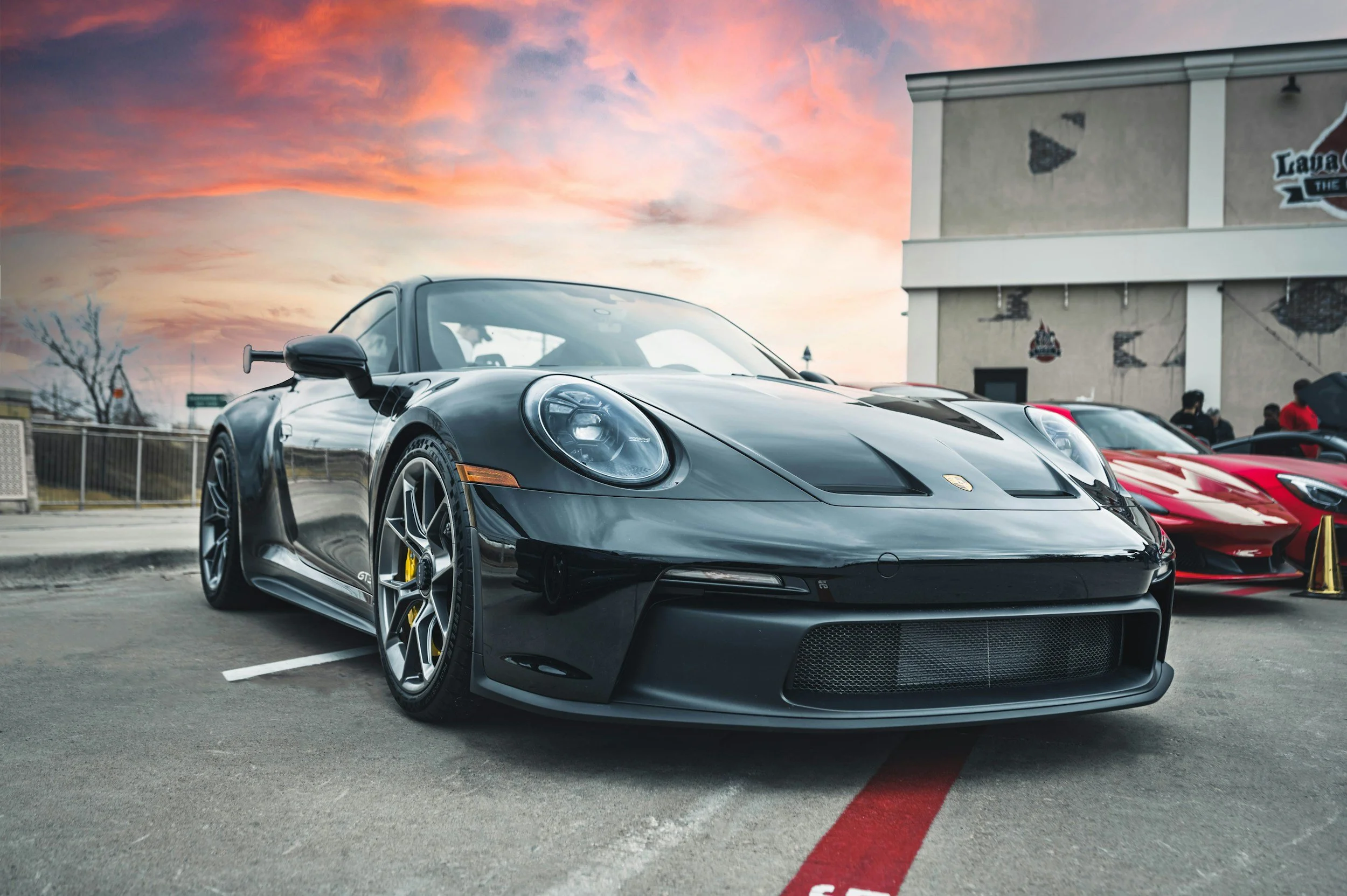 A black Porsche sports car parked outdoors at sunset with a pink and purple sky. Other cars and a building with a mural are visible in the background.