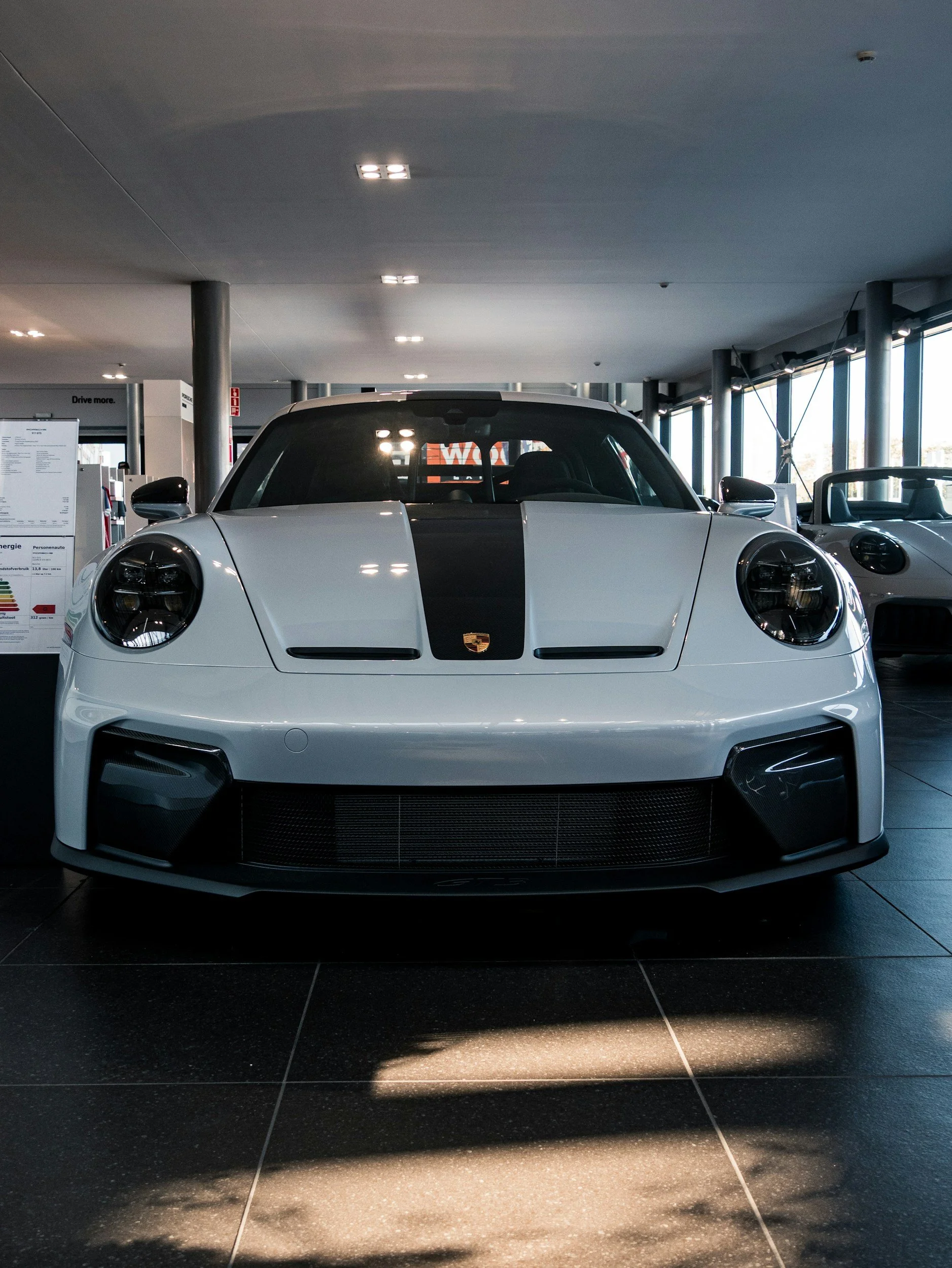 White sports car with a black stripe down the center of the hood, parked indoors in a showroom or garage. The car has a Porsche emblem on the front and is surrounded by large windows, with reflections of ceiling lights visible on its windshield.