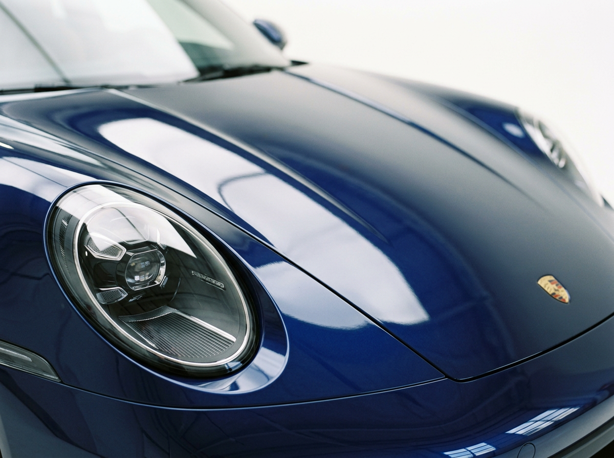 Close-up view of a sleek, dark blue Porsche sports car, highlighting the front left headlight and part of the hood with the Porsche emblem.
