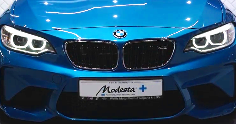 Front view of a blue BMW car with illuminated headlights and a black grille, showing a European license plate with the word 'Modesta' and a blue plus sign.