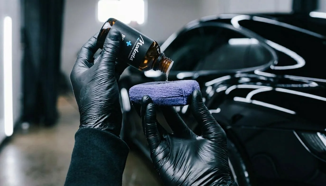 Person wearing black gloves pouring liquid from a brown bottle onto a purple cloth in front of a black car.