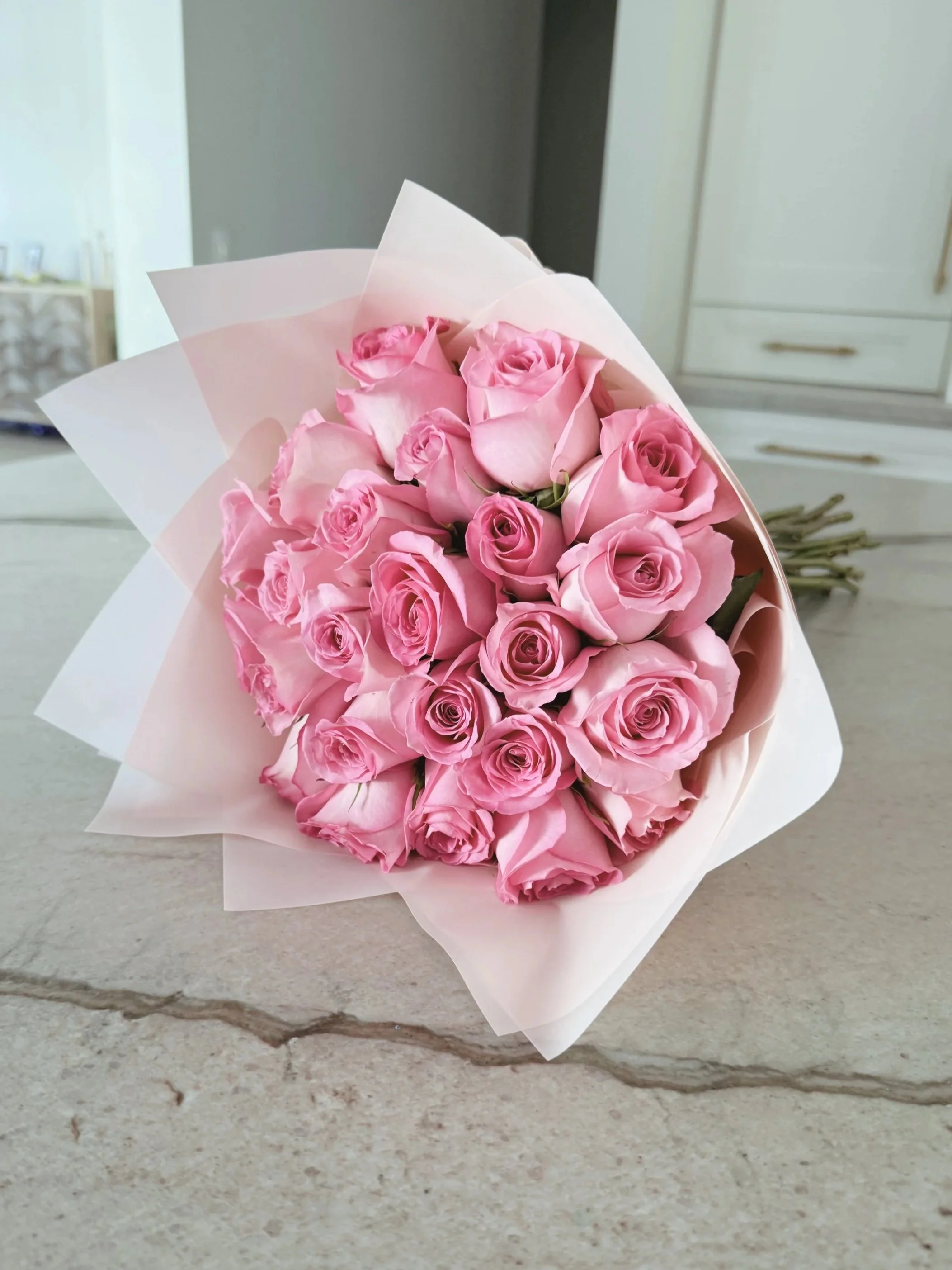 A bouquet of pink roses wrapped in white paper, placed on a beige countertop.