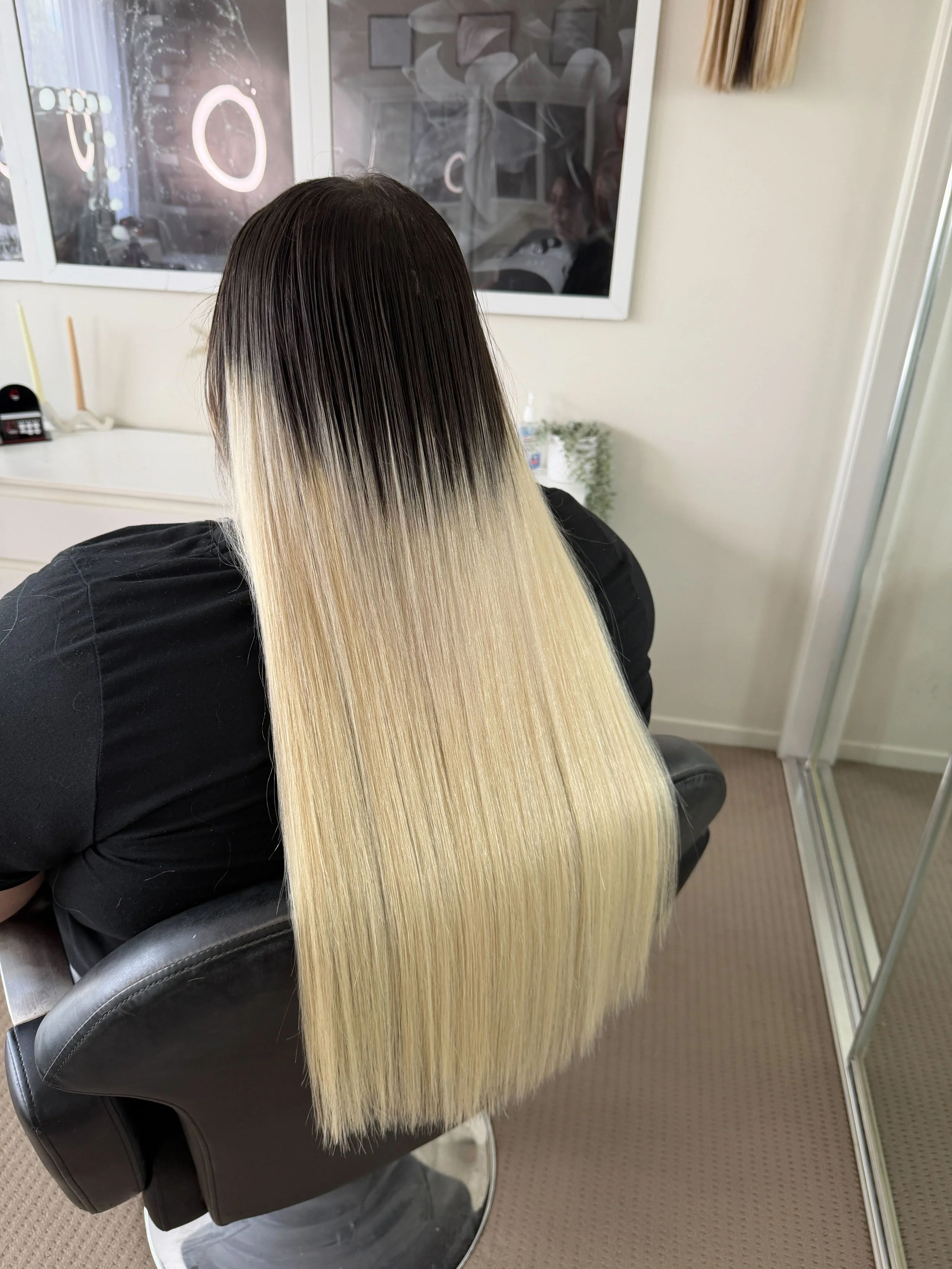 Back of a woman with long, blonde ombre hair, sitting in a salon chair in front of a mirror, getting her hair straightened or styled.