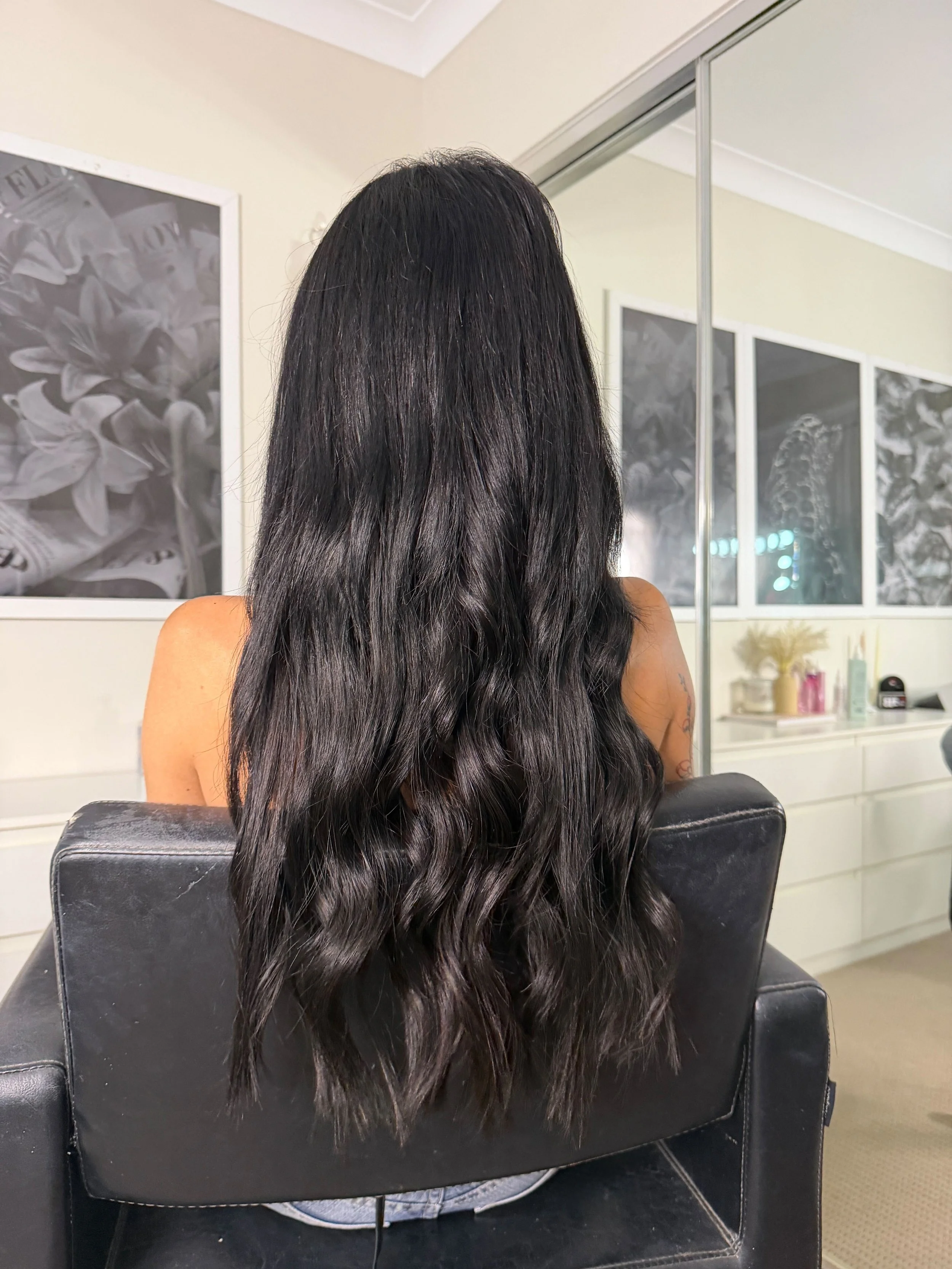 Back view of a person with long, wavy black hair sitting in a salon chair, facing away, with a large mirror and salon supplies visible in the background.