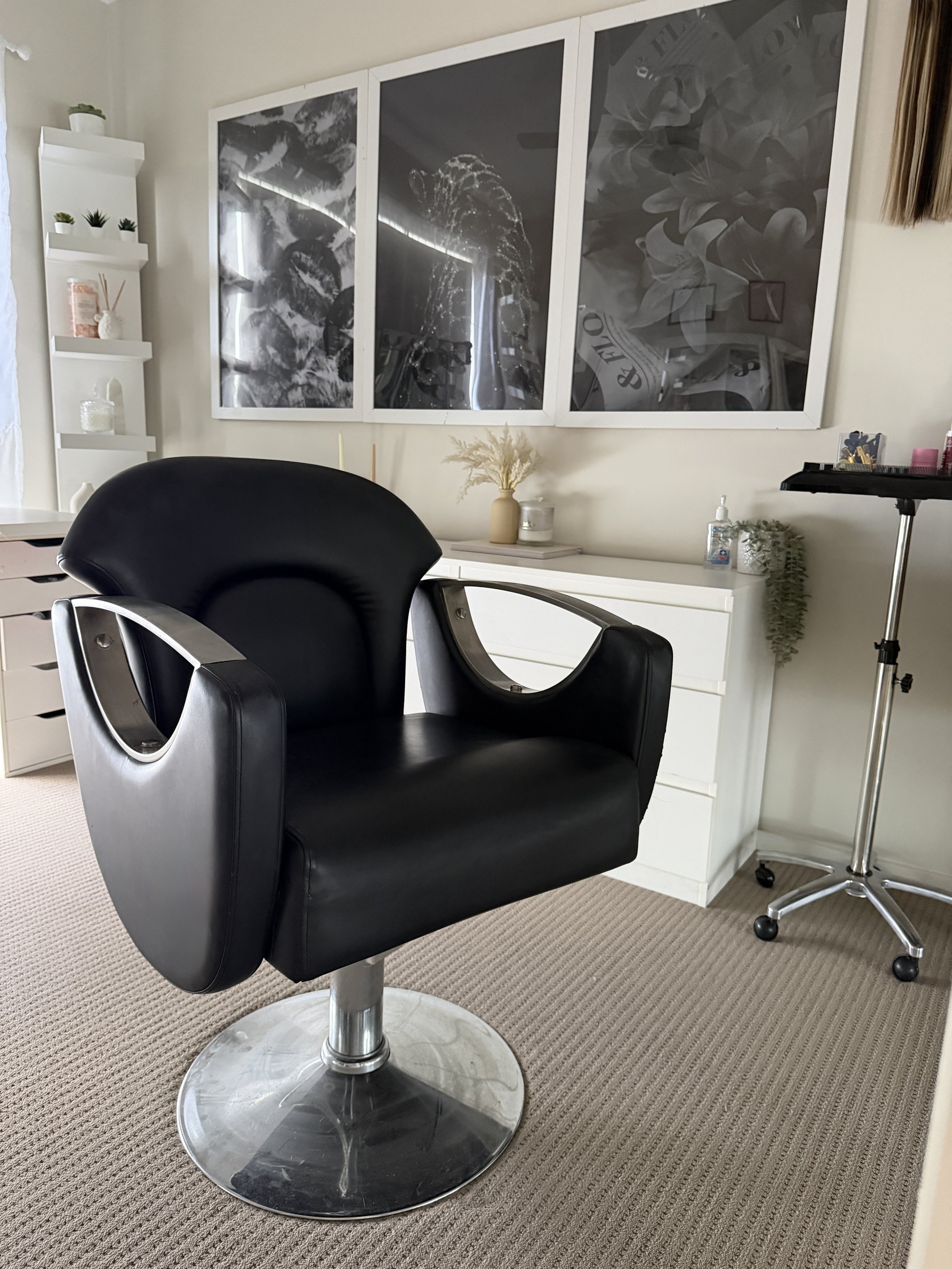 A black salon chair with a circular metal base in a room with beige carpet and white walls. In the background, there is a white dresser, a large white cabinet with decorative items on top, and three framed black and white artworks on the wall. There is also a small white table with a vase and decorative items, and a rolling cart with skincare products and tools.