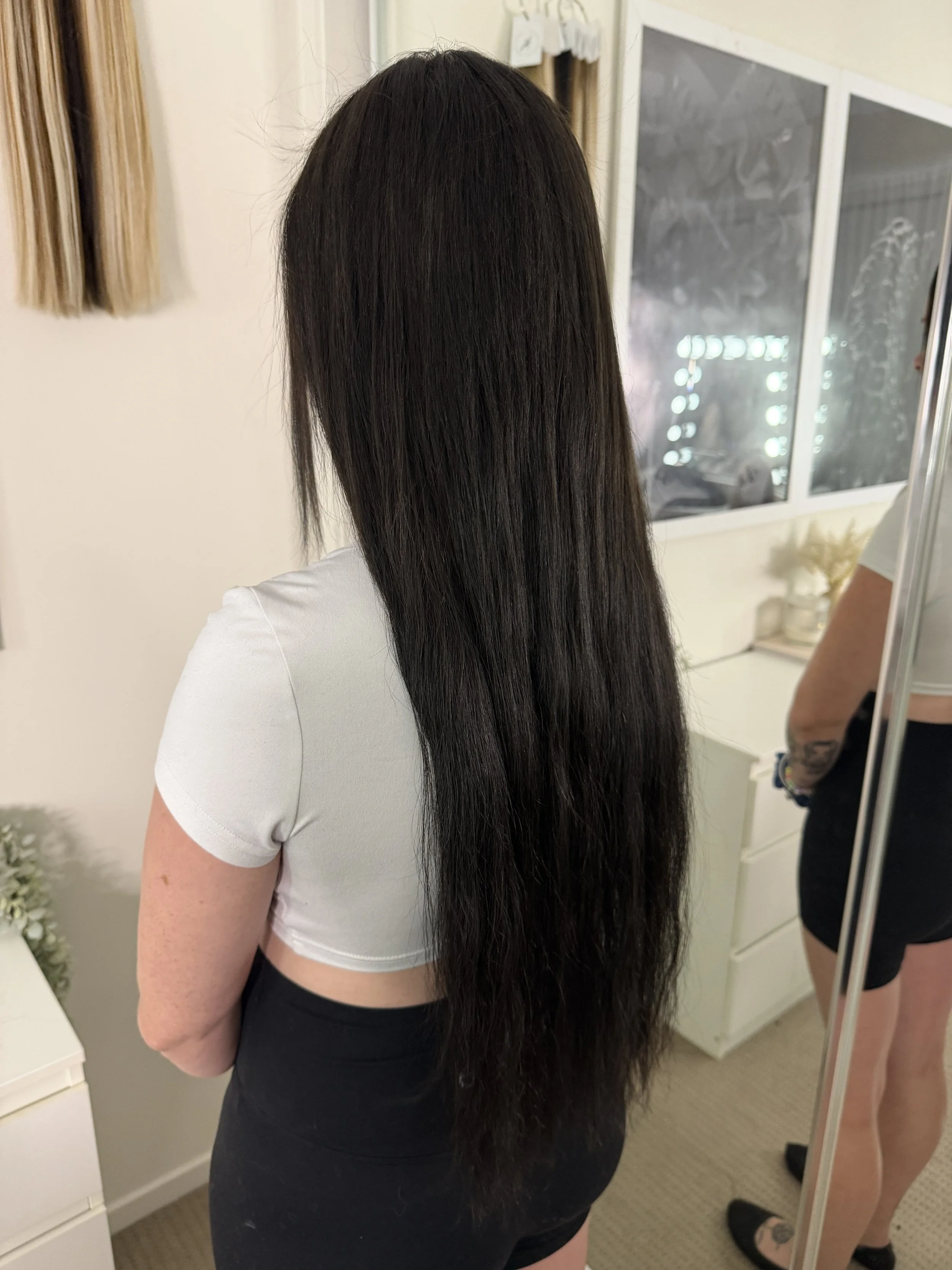 A woman with long, straight, dark hair standing in front of a mirror, showing her hair from the back. She is wearing a white top and black shorts, and there is another person with tattoos on their arm and legs visible in the background.