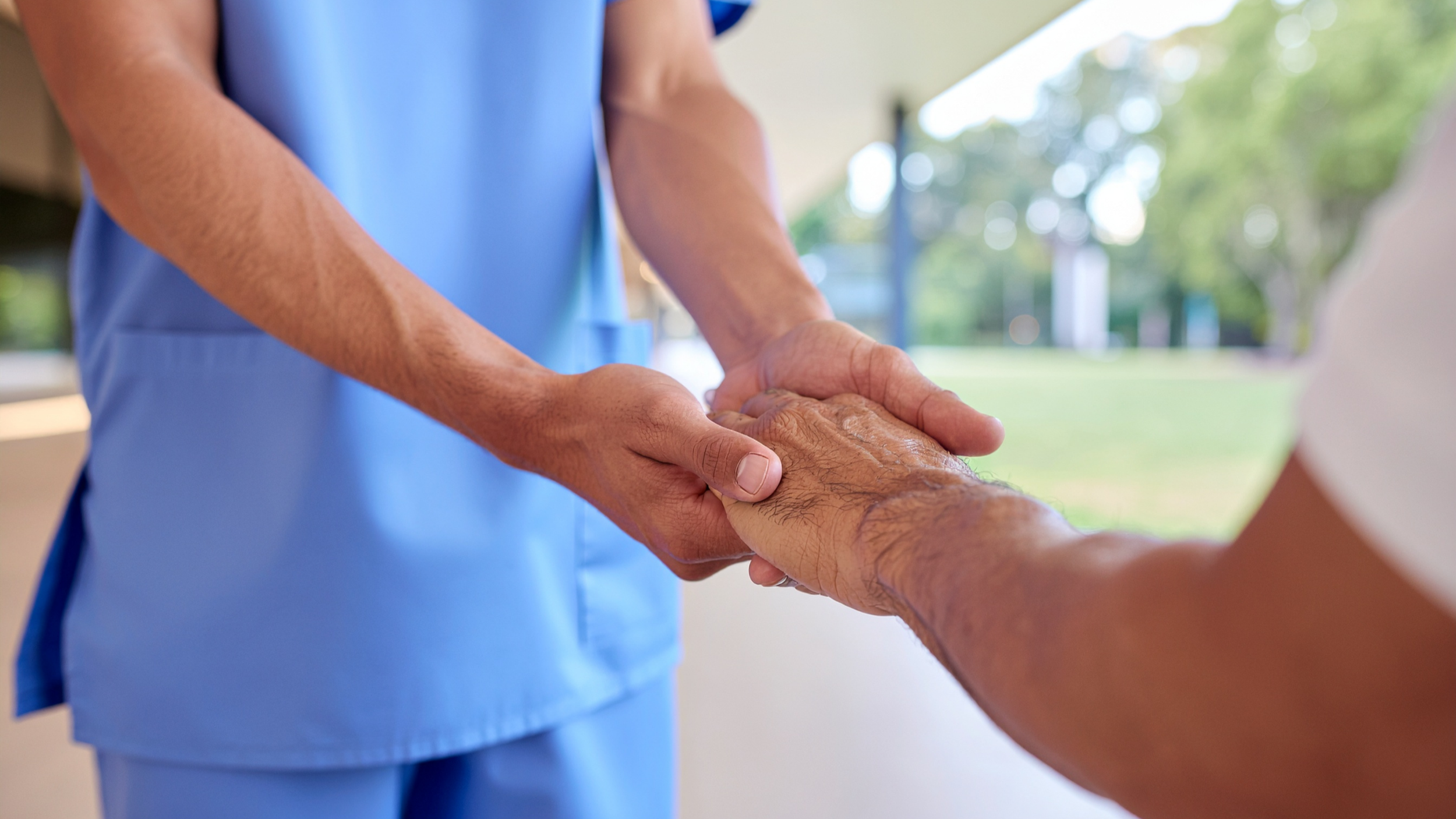 A healthcare worker in a blue uniform holding a patient's hand outdoors with greenery in the background.