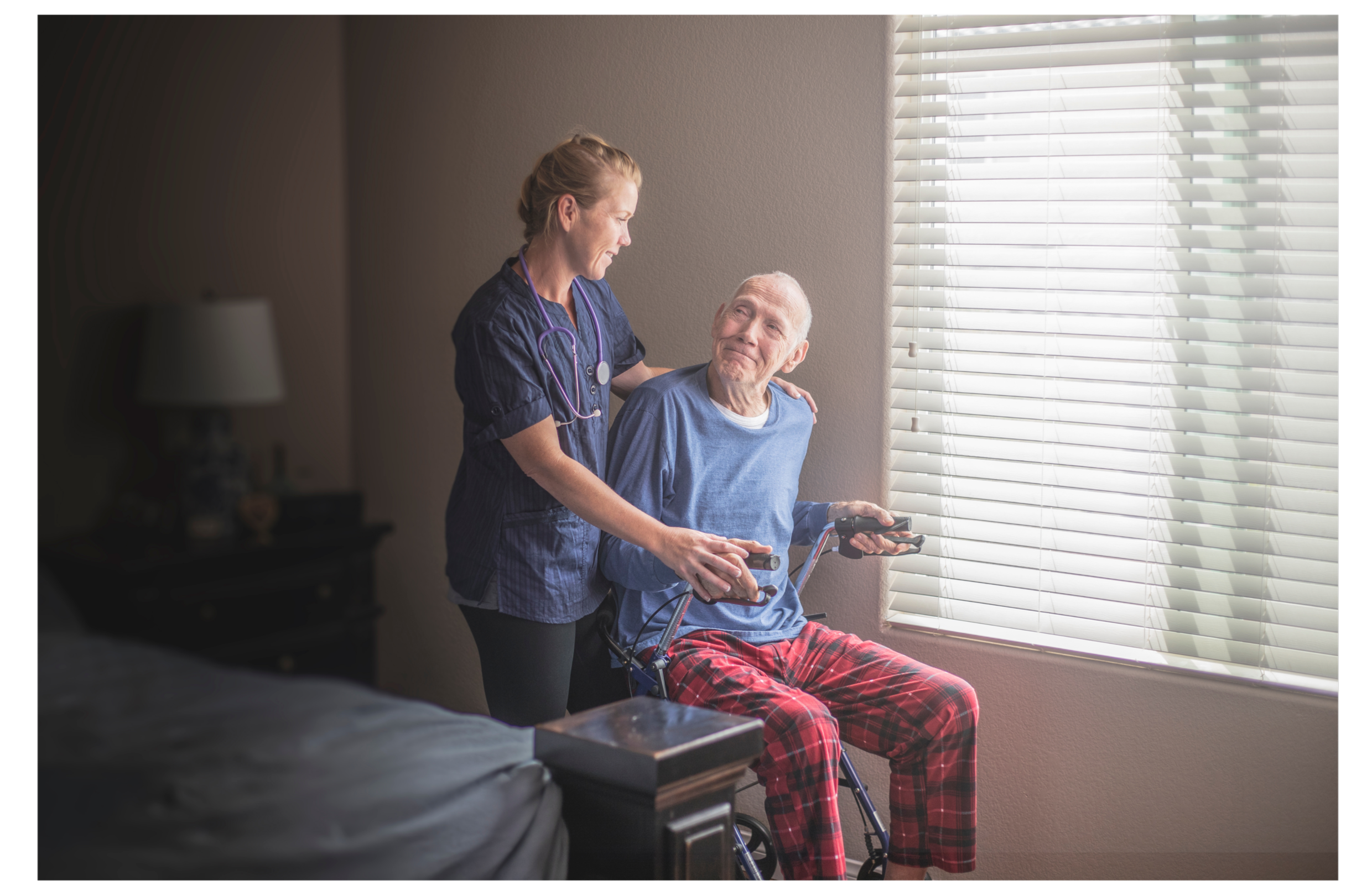 Nurse assisting elderly man in a motorized wheelchair, near a window with blinds, in a home setting.