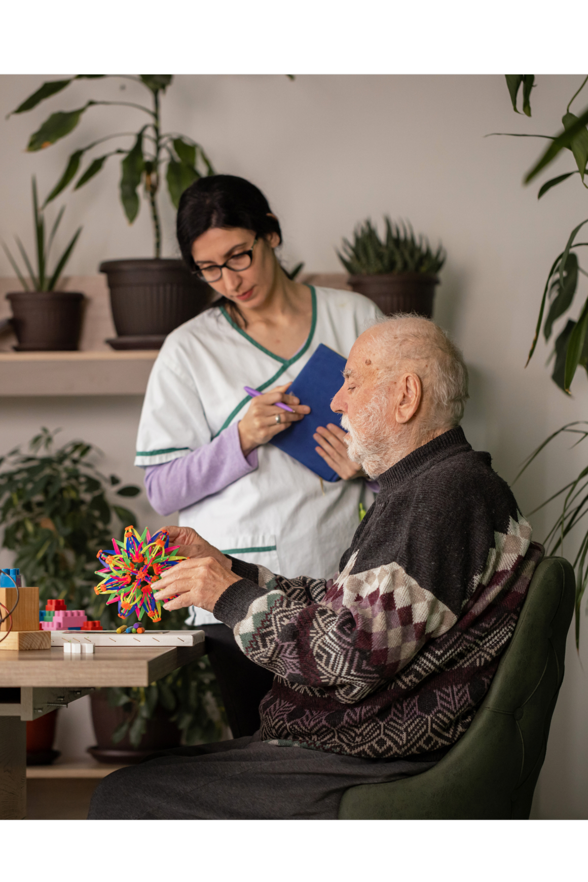 An elderly man with a white beard playing with a colorful 3D geometric brain model at a table, while a caregiver and a healthcare worker observe and note in a clipboard, in a room with plants.