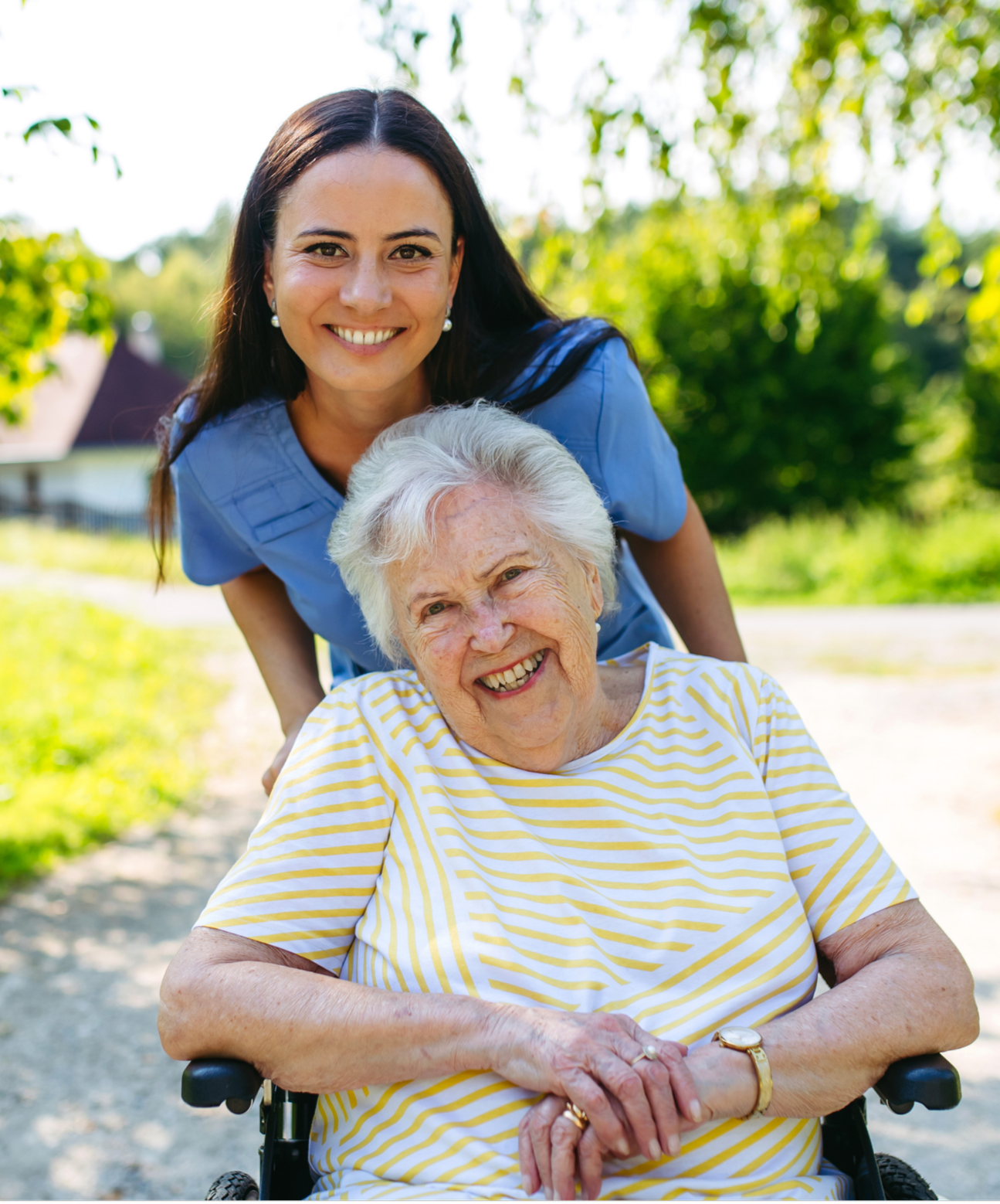 A young woman in a blue uniform leaning over and smiling with an elderly woman in a yellow and white striped shirt sitting in a wheelchair outdoors, with green trees and a bright sky in the background.