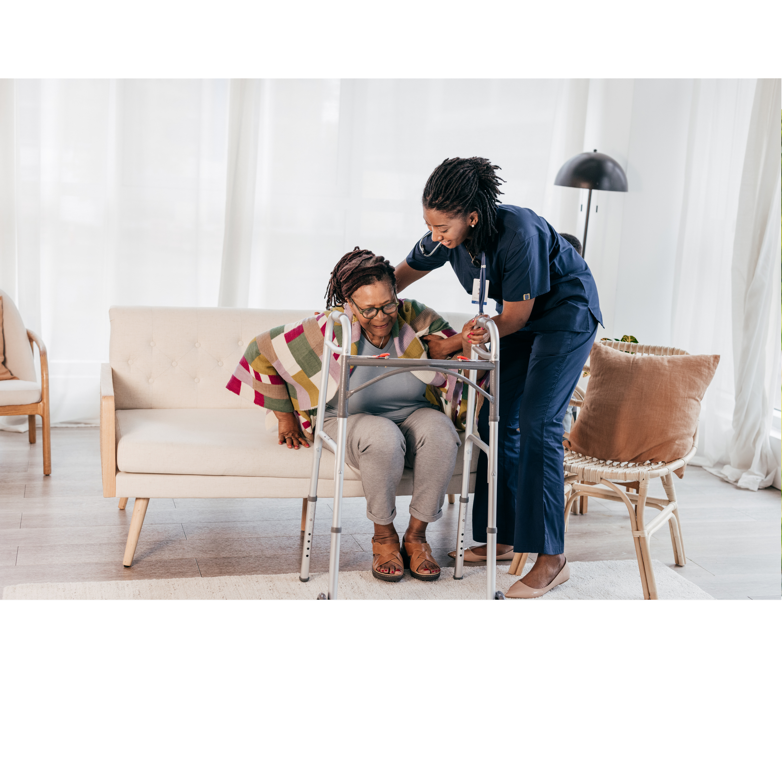 A nurse helping an elderly woman with a walker in a well-lit room with white curtains. The nurse is smiling and assisting as the woman, seated on a sofa, wears glasses and a colorful striped shawl.