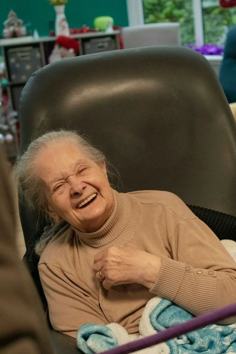An elderly woman with gray hair is laughing joyfully while sitting in a chair, covered with a blanket, in a room with holiday decorations in the background.