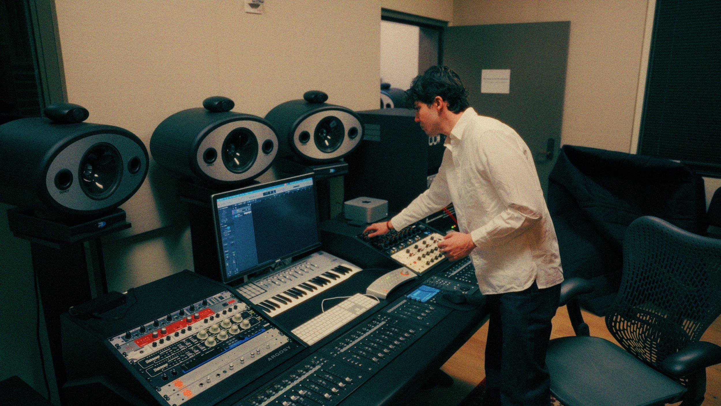 A man in a white shirt working with music production equipment in a recording studio, surrounded by speakers and electronic music gear.
