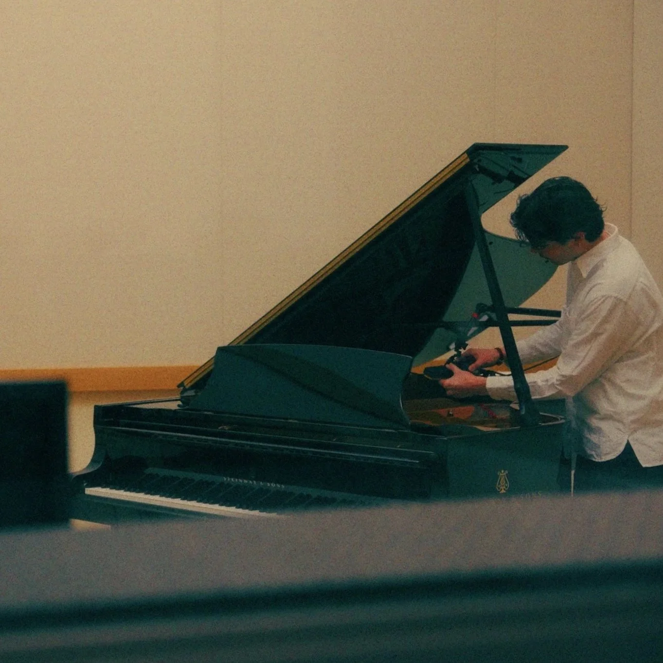 A man with curly dark hair in a white shirt is standing next to a black grand piano, adjusting some equipment or settings.