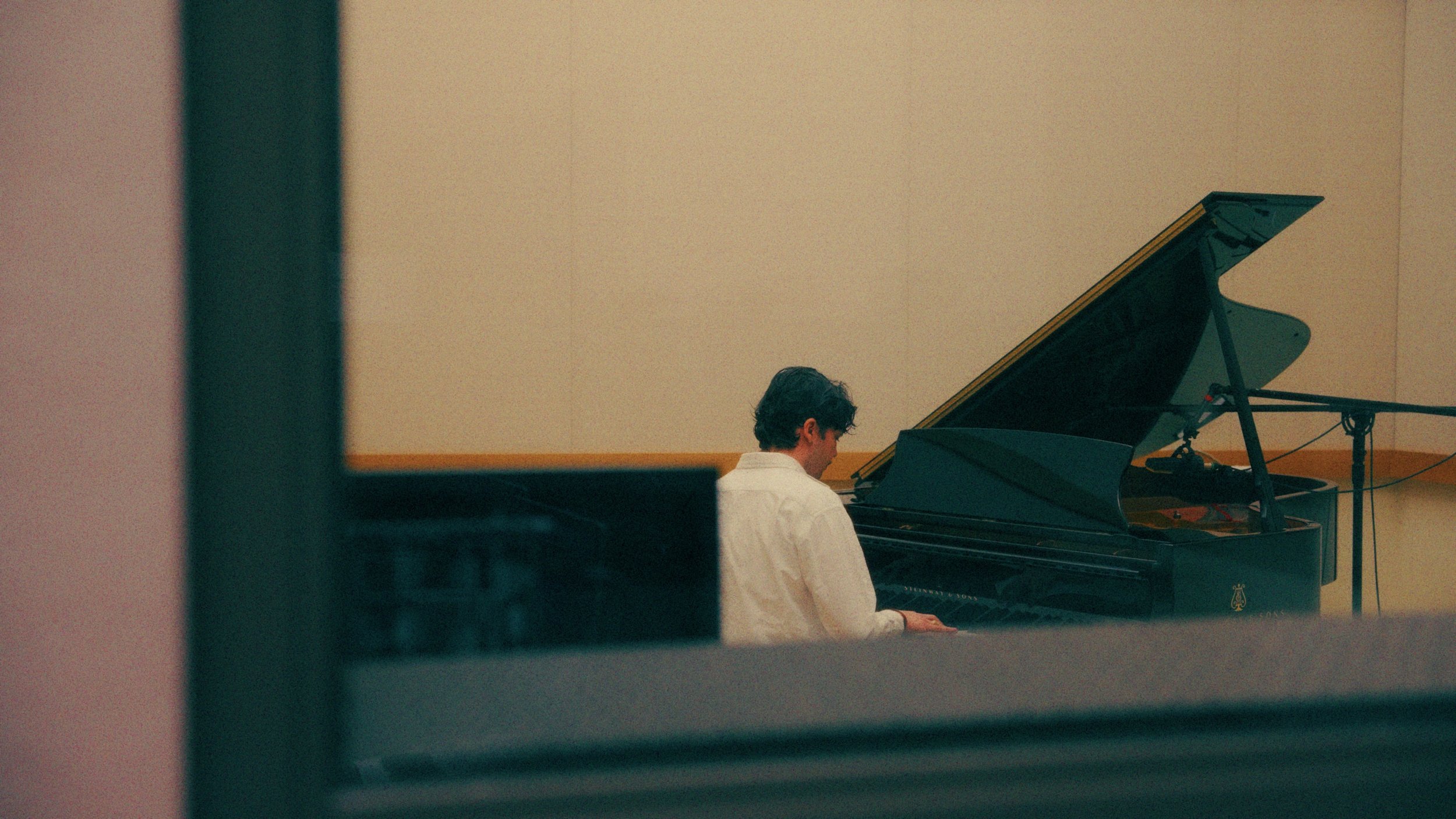 A man with dark, curly hair wearing a white shirt is playing a grand piano in a room with plain beige walls. The image is framed as if viewed through a partially opened door or window.