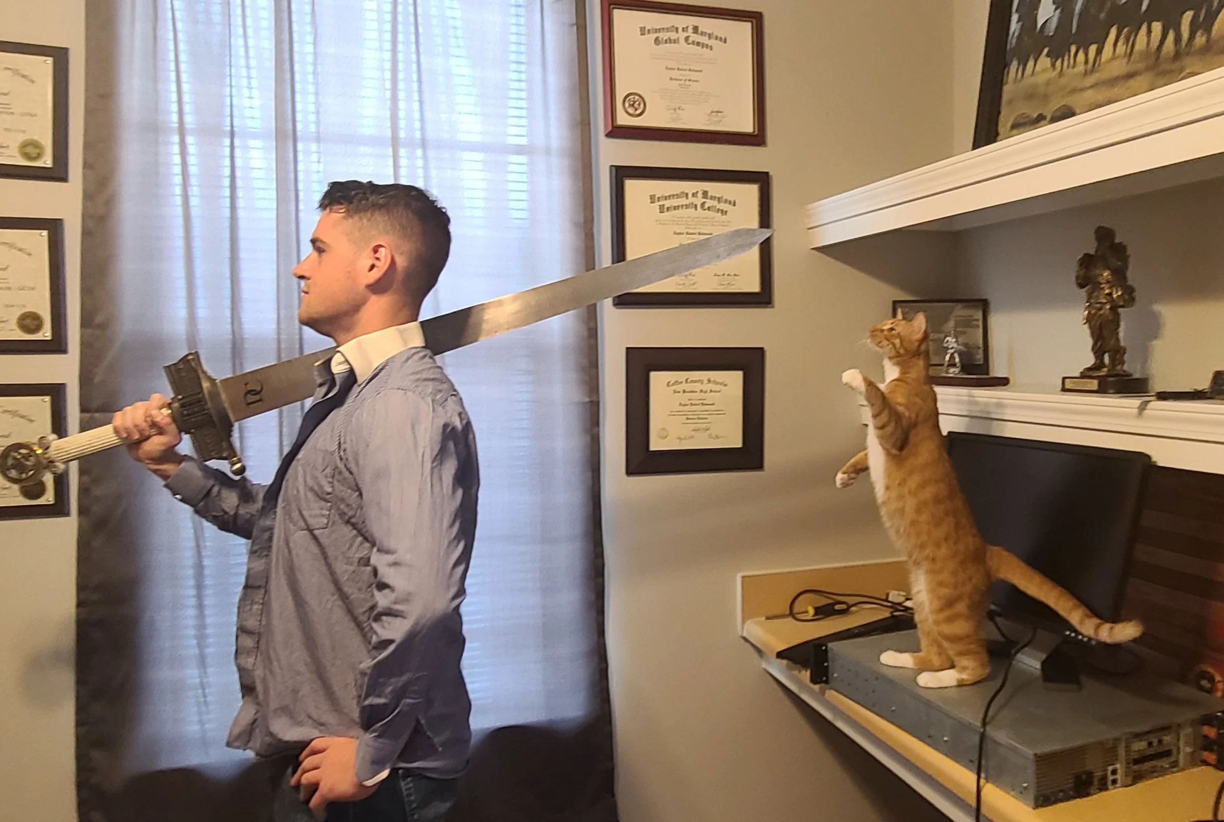 Author Eadi Q. Blagit, wearing a gray shirt, poses with a large sword while his orange cat, Garfield, stands on his hind legs beside him atop a desk in his home office.