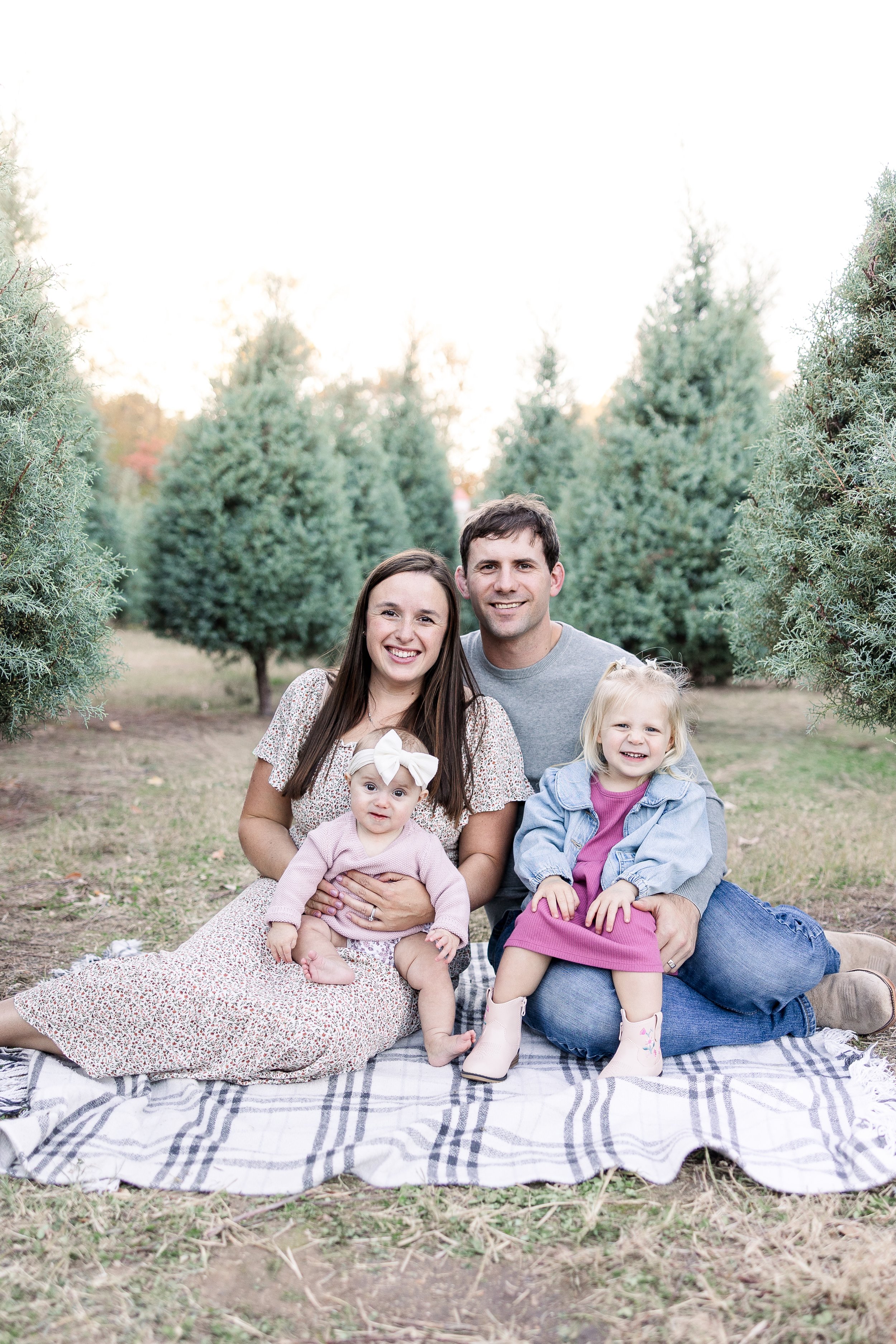 A family of five sitting on a blanket outdoors, surrounded by evergreen trees, smiling at the camera.